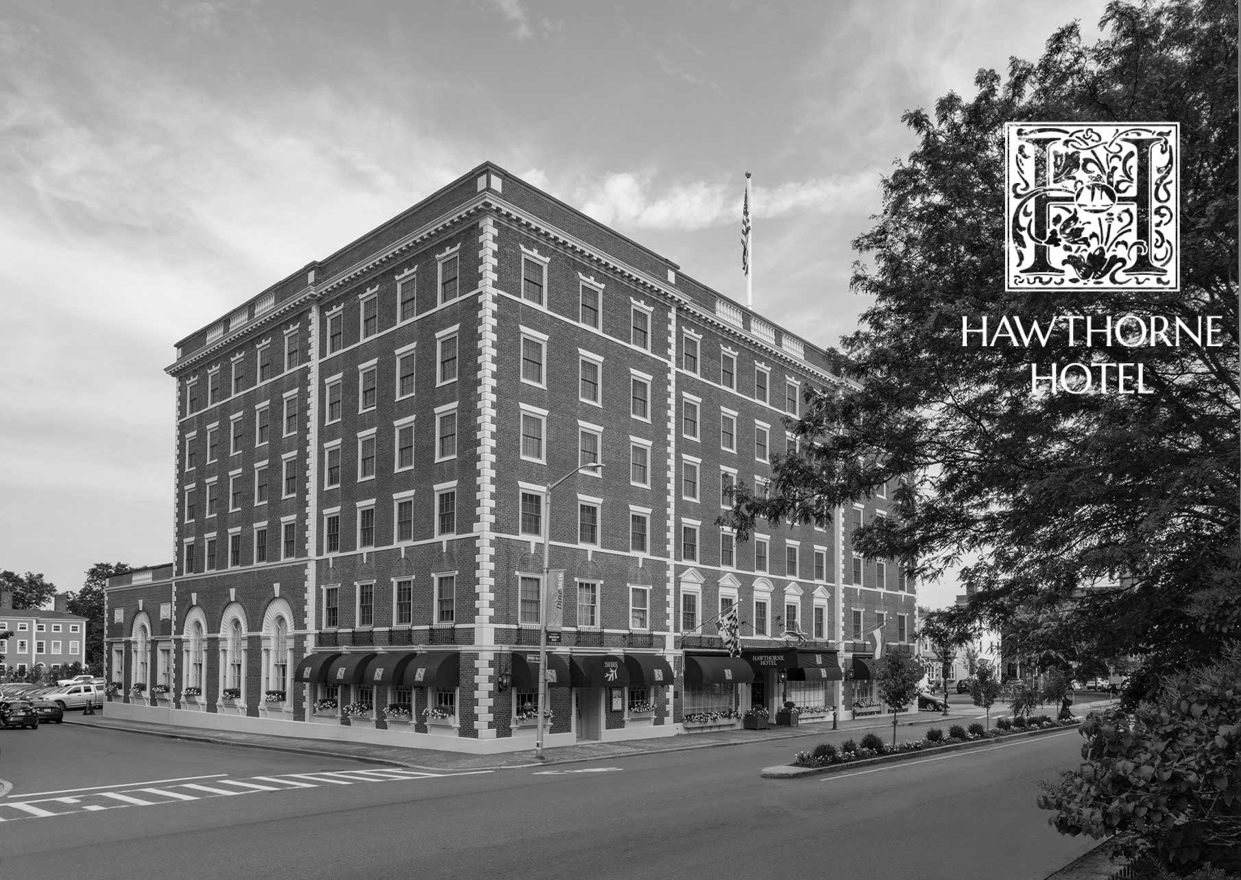 Black and white photo of the Hawthorne Hotel, a multi-story brick building with arched windows and striped awnings on the ground level, located on a street corner with trees and parked cars nearby.