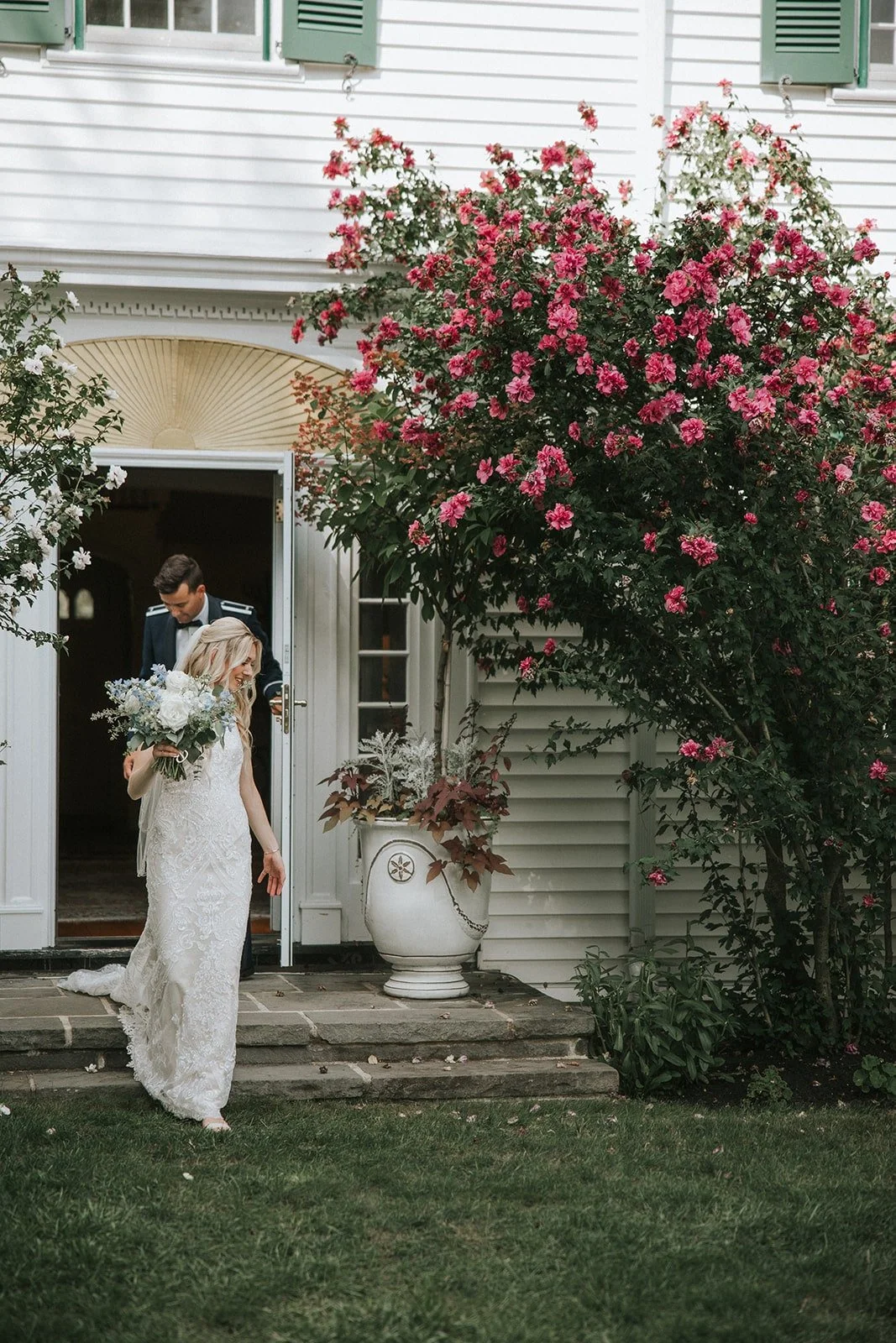 A bride and groom exiting a house, with the bride holding a bouquet of white and blue flowers, surrounded by pink flowering bushes and a white house exterior.