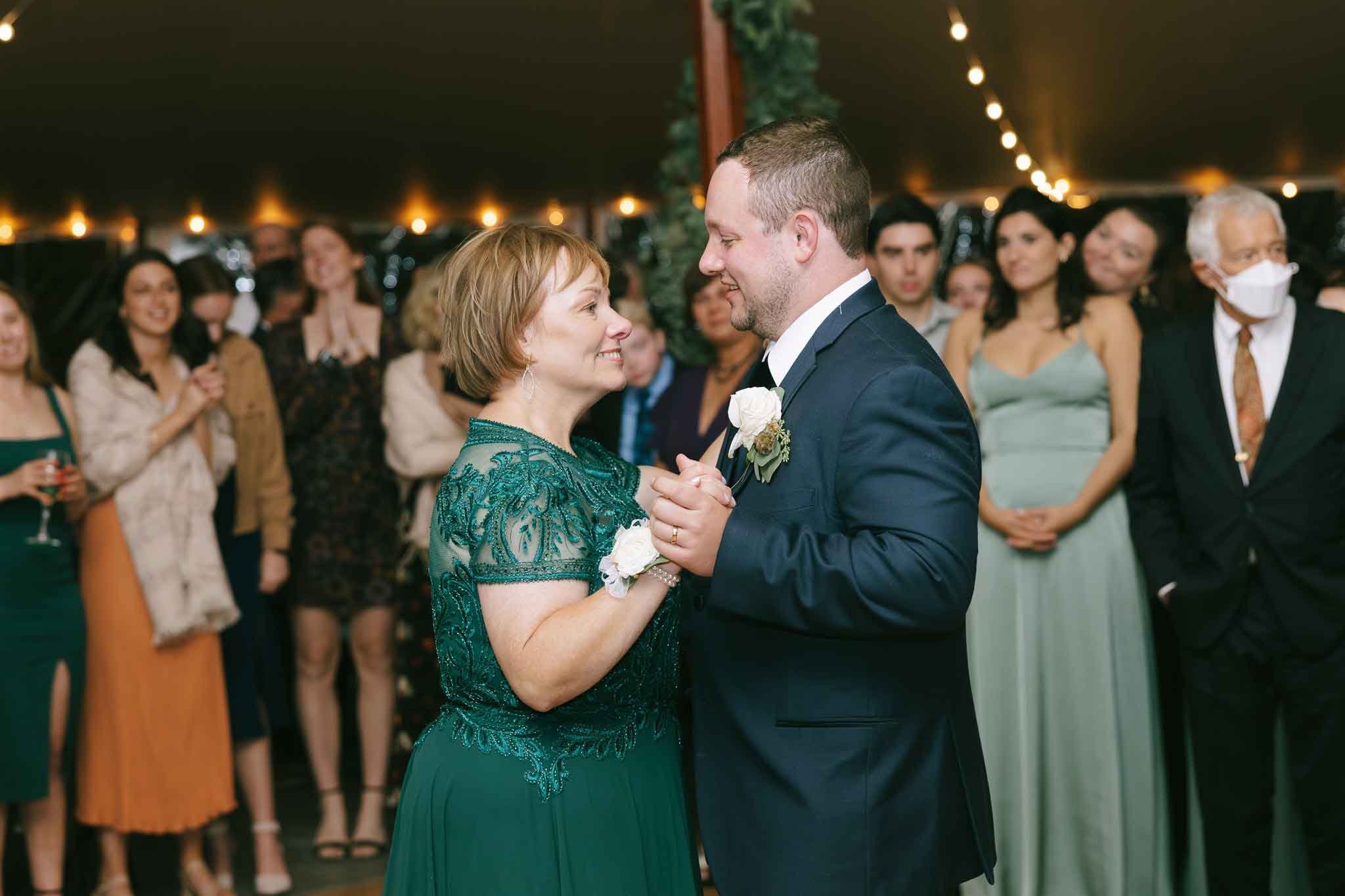 A mother and son dance at a wedding reception, surrounded by friends and family under a tent with string lights.