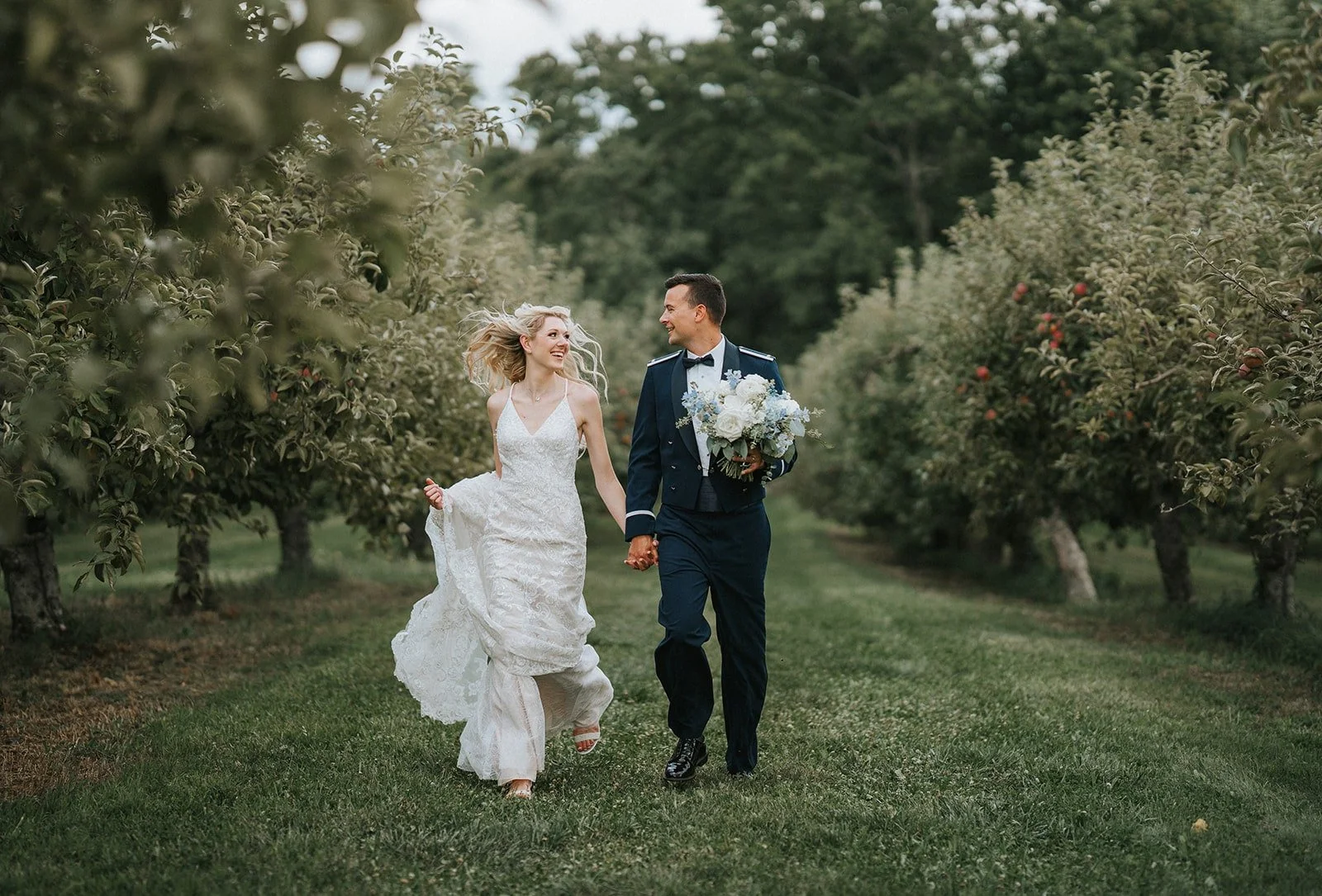 A bride and groom walking hand in hand through a lush orchard, smiling and looking at each other. The bride is wearing a white lace wedding dress and the groom is dressed in a navy military uniform, holding a bouquet of white flowers.