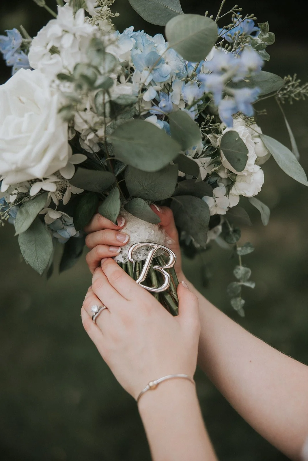 A person holding a bouquet of white and blue flowers with green leaves, the bouquet is wrapped with a ribbon that has a silver letter 'B', and they are wearing a ring and a bracelet on their hands.