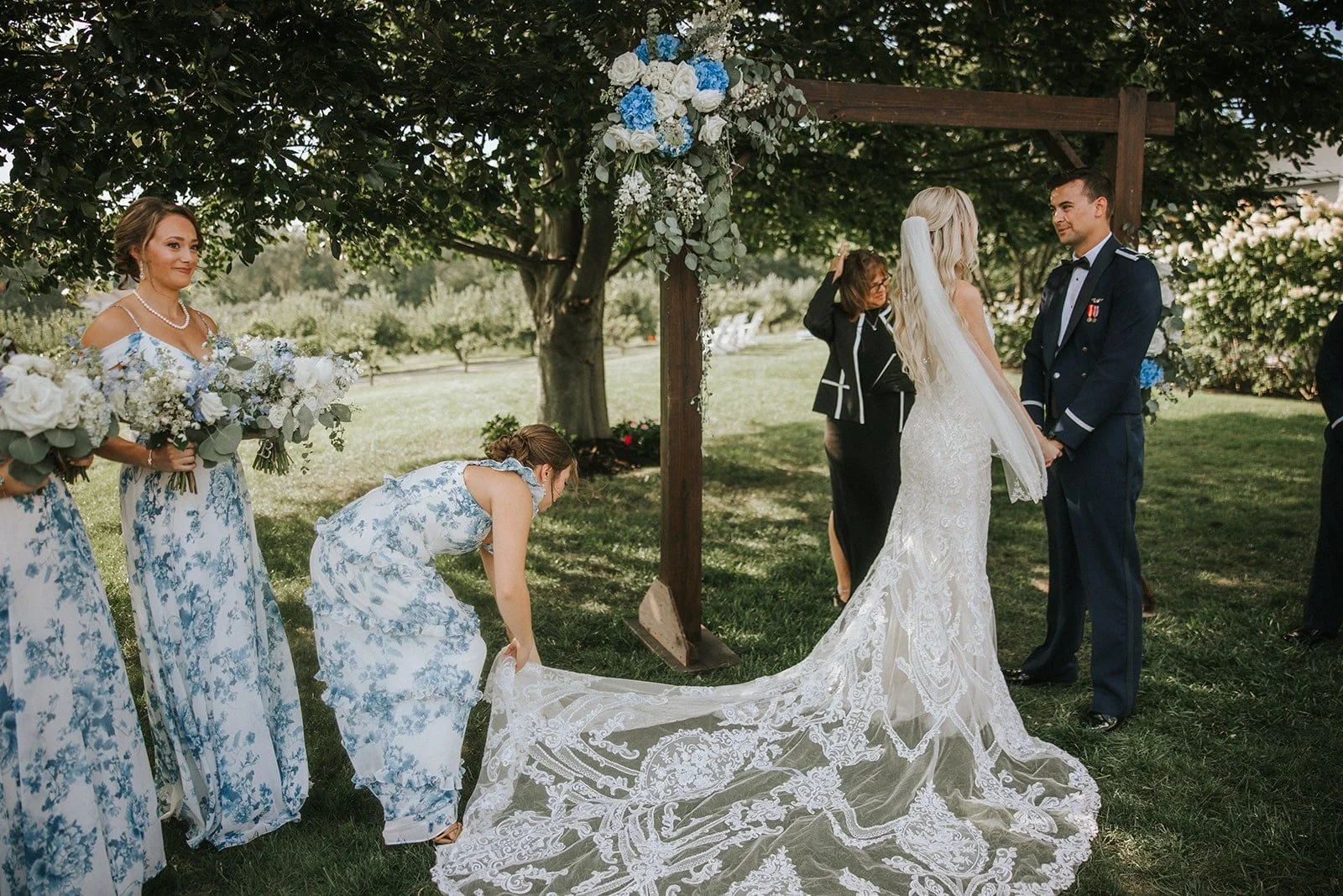 A wedding ceremony happening outdoors under a tree, with a bride in a white lace wedding gown and veil, and a groom in a navy uniform. Bridesmaids in matching blue and white floral dresses hold bouquets, and one bridesmaid adjusts the bride's train. 