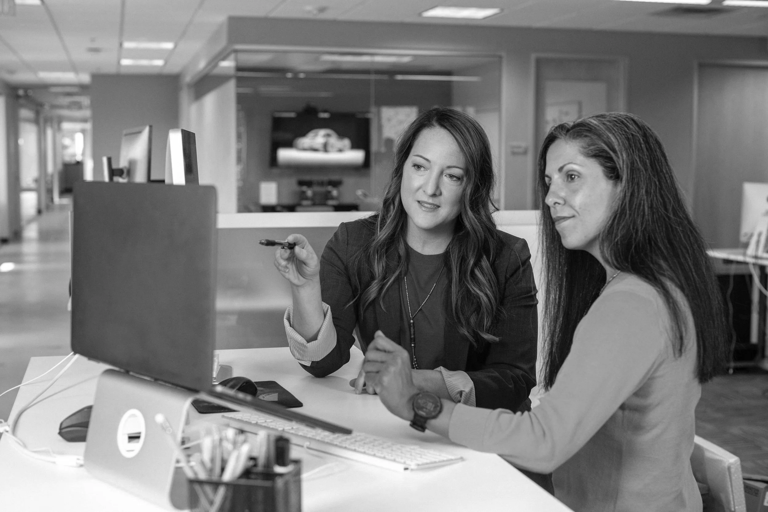 Two women having a discussion at a desk in an office environment, looking at a computer monitor, with a background of office furniture and decor.
