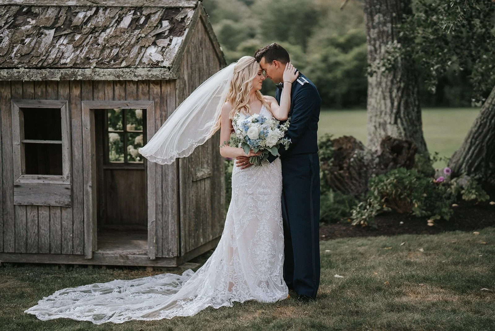 A bride and groom share a kiss outdoors, with the bride holding a bouquet of white and blue flowers, next to an old wooden shed and surrounded by trees.