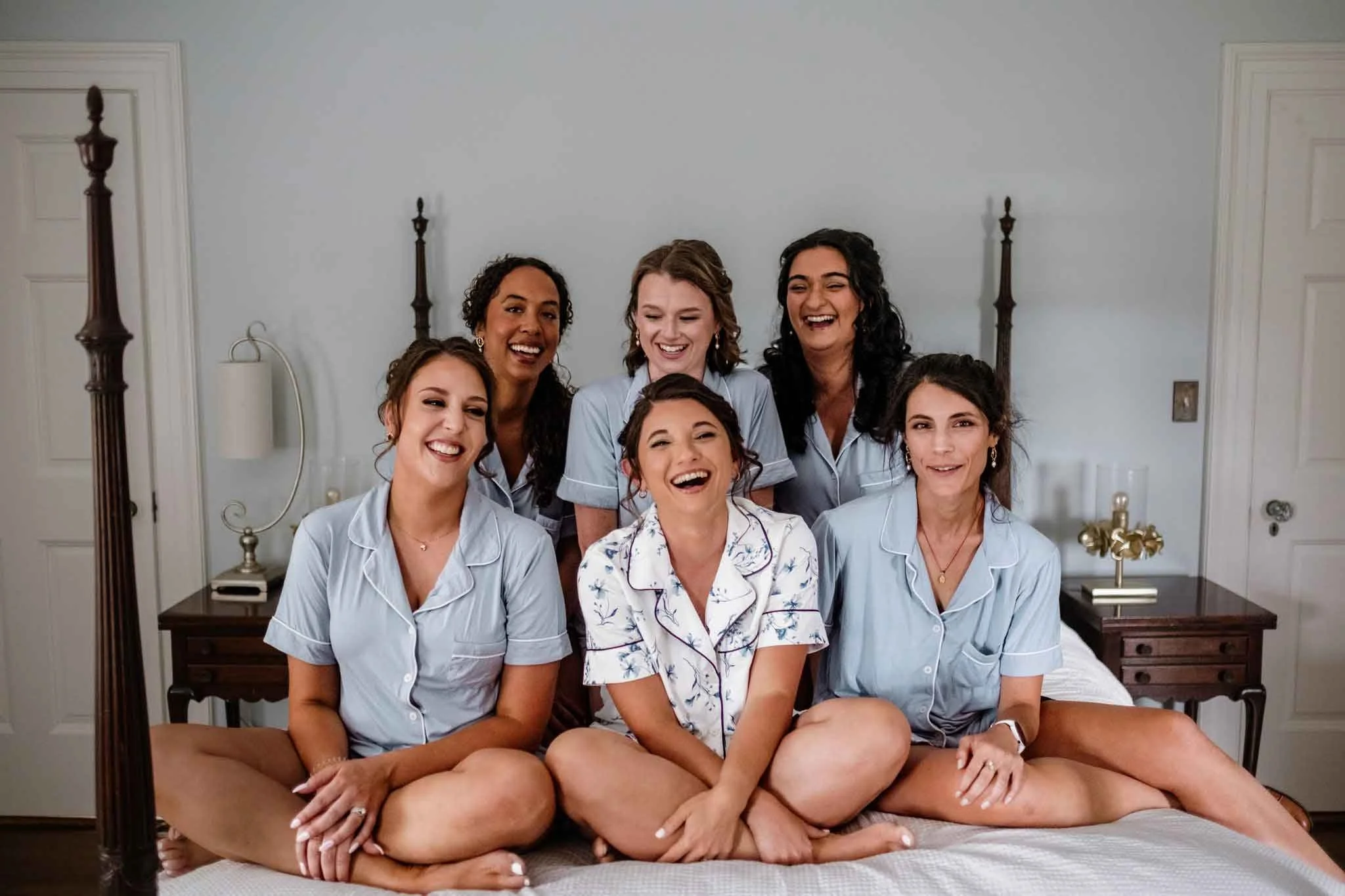 Seven women sitting on a bed in pajamas, smiling and laughing during a joyful moment in a bedroom.