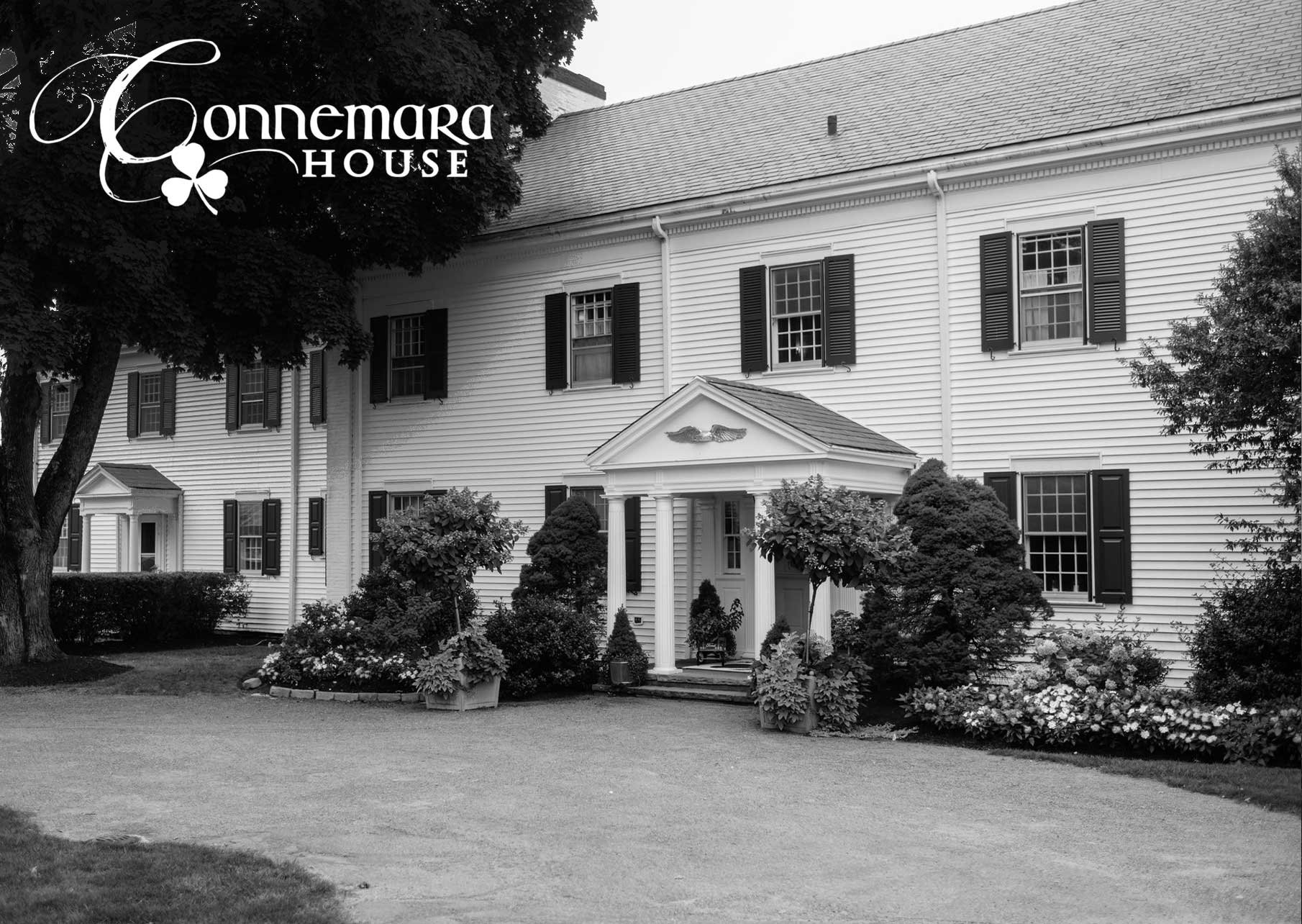 A black and white photo of the Connemara House, a large white building with black shutters, a small porch with columns, and an arrangement of trees and bushes in the foreground.