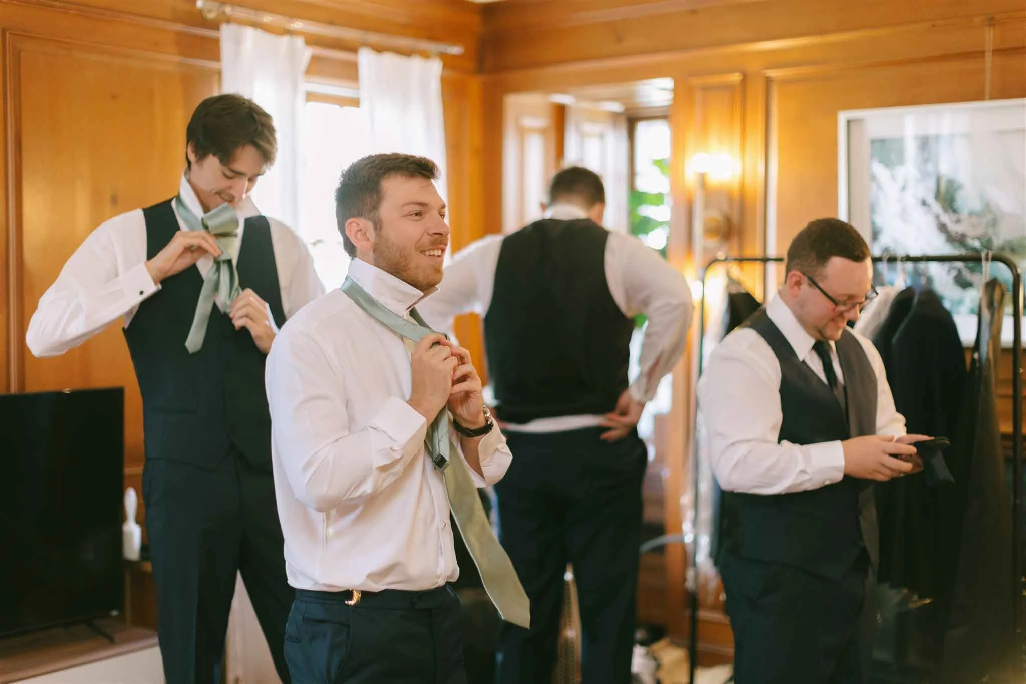 Four men in formal attire in a wooden-paneled room preparing for an event, with two tying ties, one using a phone, and one adjusting his waistband.