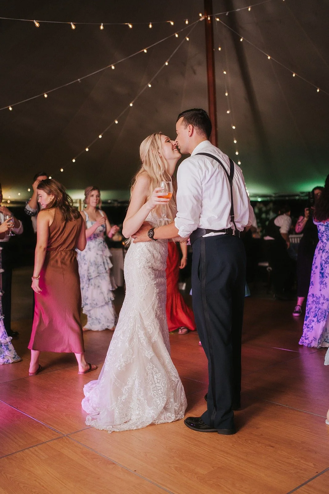 A bride and groom dancing closely at their wedding reception inside a decorated tent with string lights, surrounded by guests.