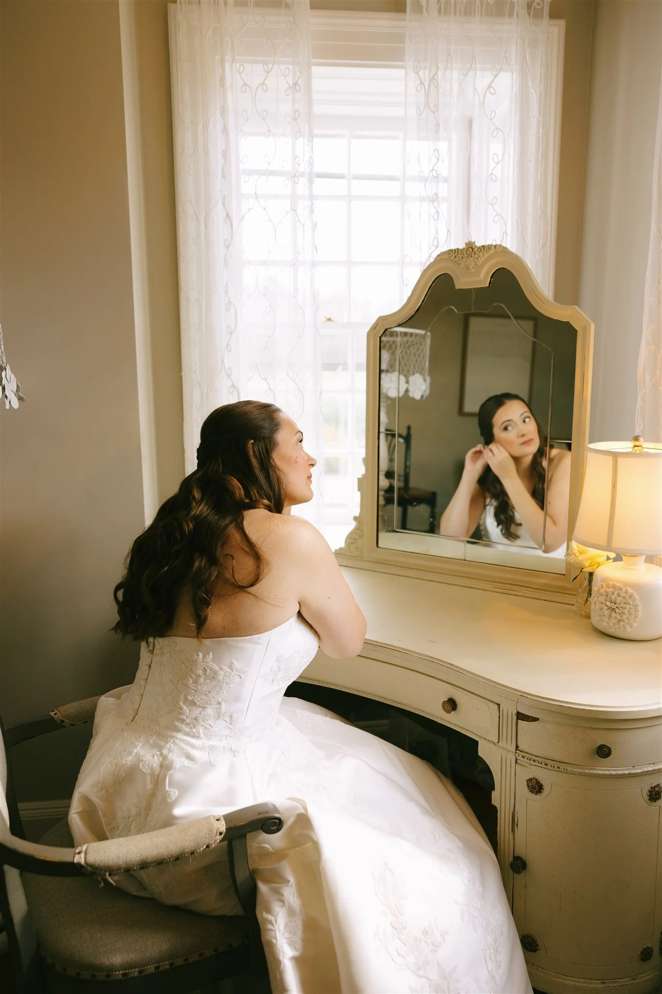 A bride in a white strapless wedding gown sitting at a vintage vanity table, looking in a mirror as she adjusts her earring.