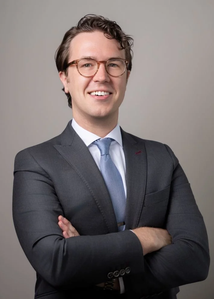 A smiling man wearing glasses, a navy suit, and a light blue tie, standing with arms crossed against a plain gray background.