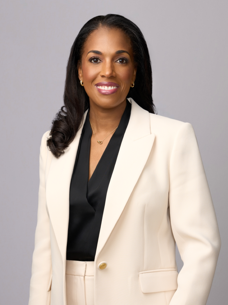 Professional woman with dark hair, wearing a white blazer over a black blouse, smiling against a plain gray background.