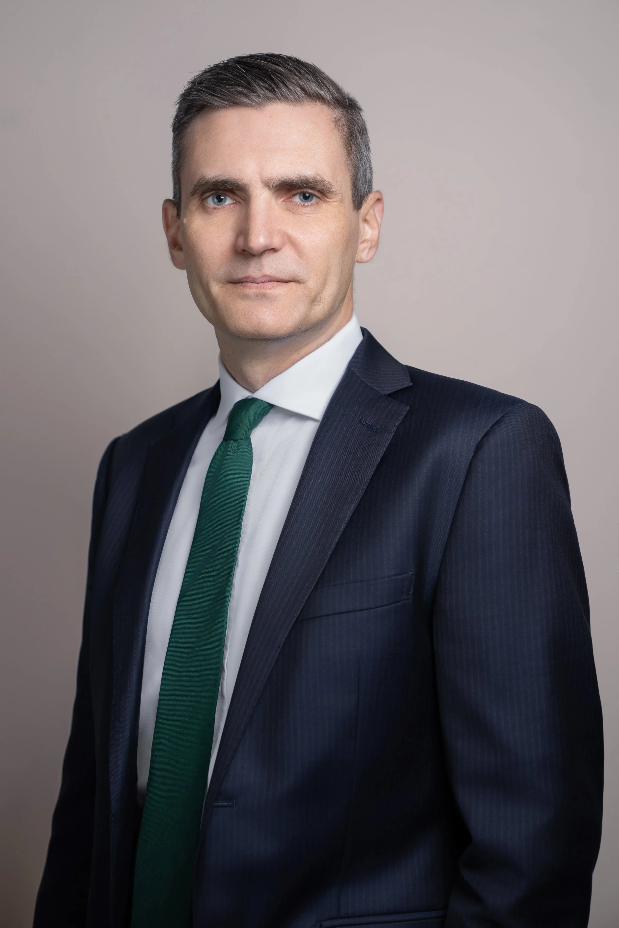 A man in a dark business suit with a white shirt and green tie, standing against a plain neutral background, looking at the camera.