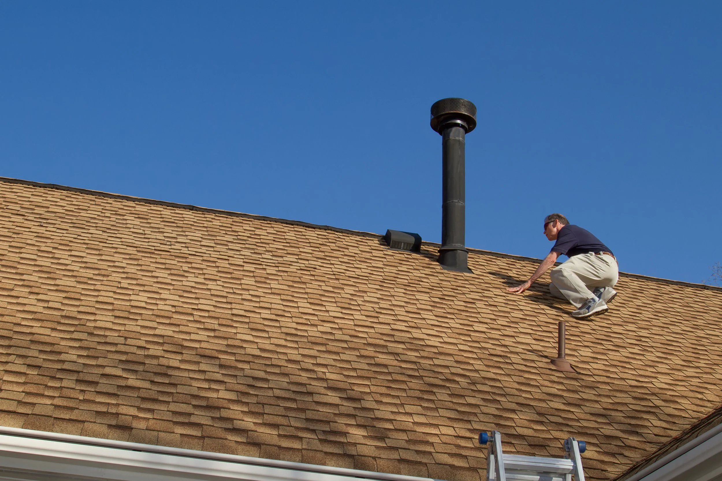 A man crouches on a residential roof inspecting shingles, with a ladder visible at the bottom and pipes and a chimney pipe on the roof, under a clear blue sky.
