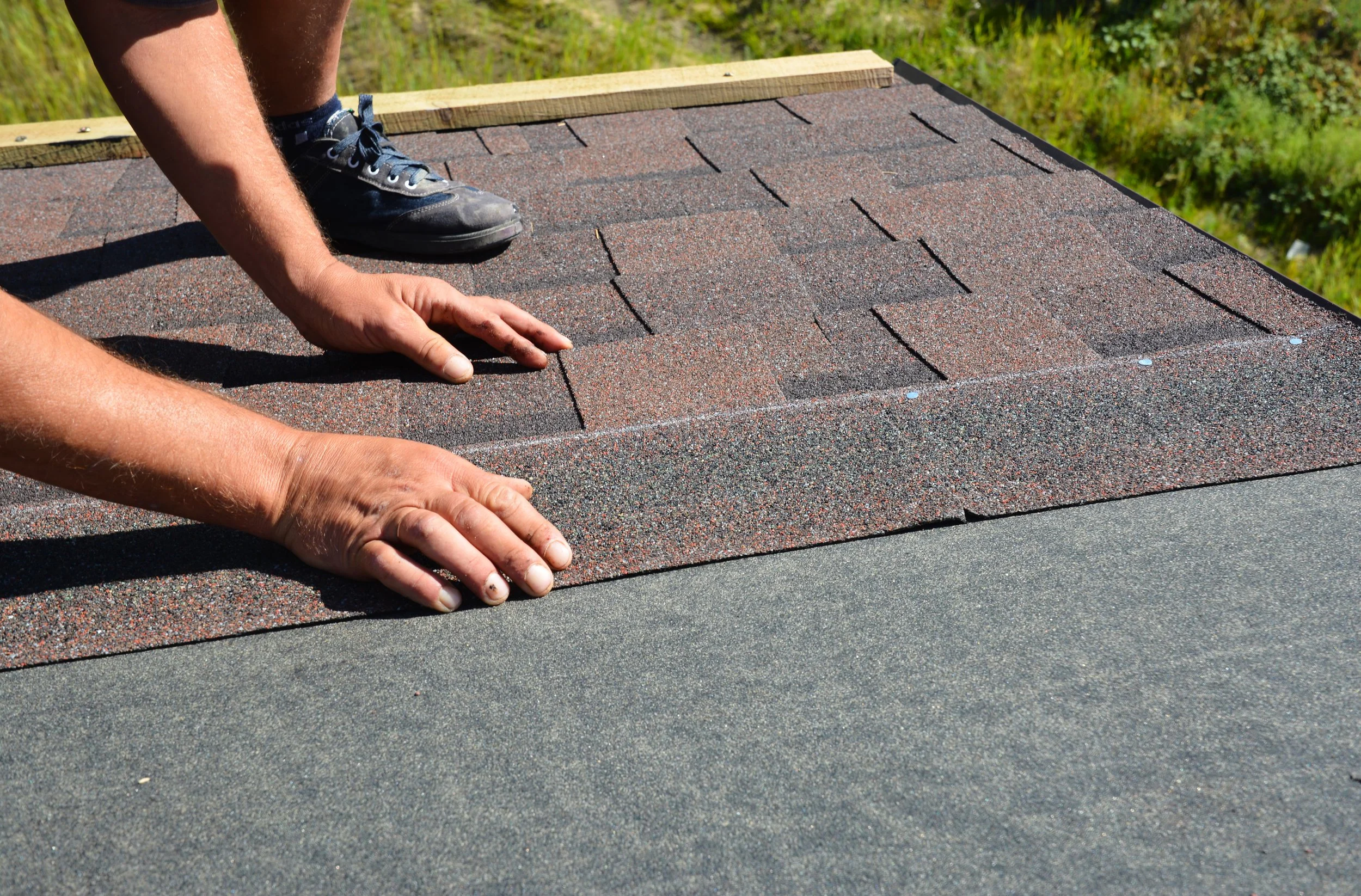 Person installing asphalt shingles on a roof, with hands and feet visible, in bright sunlight.