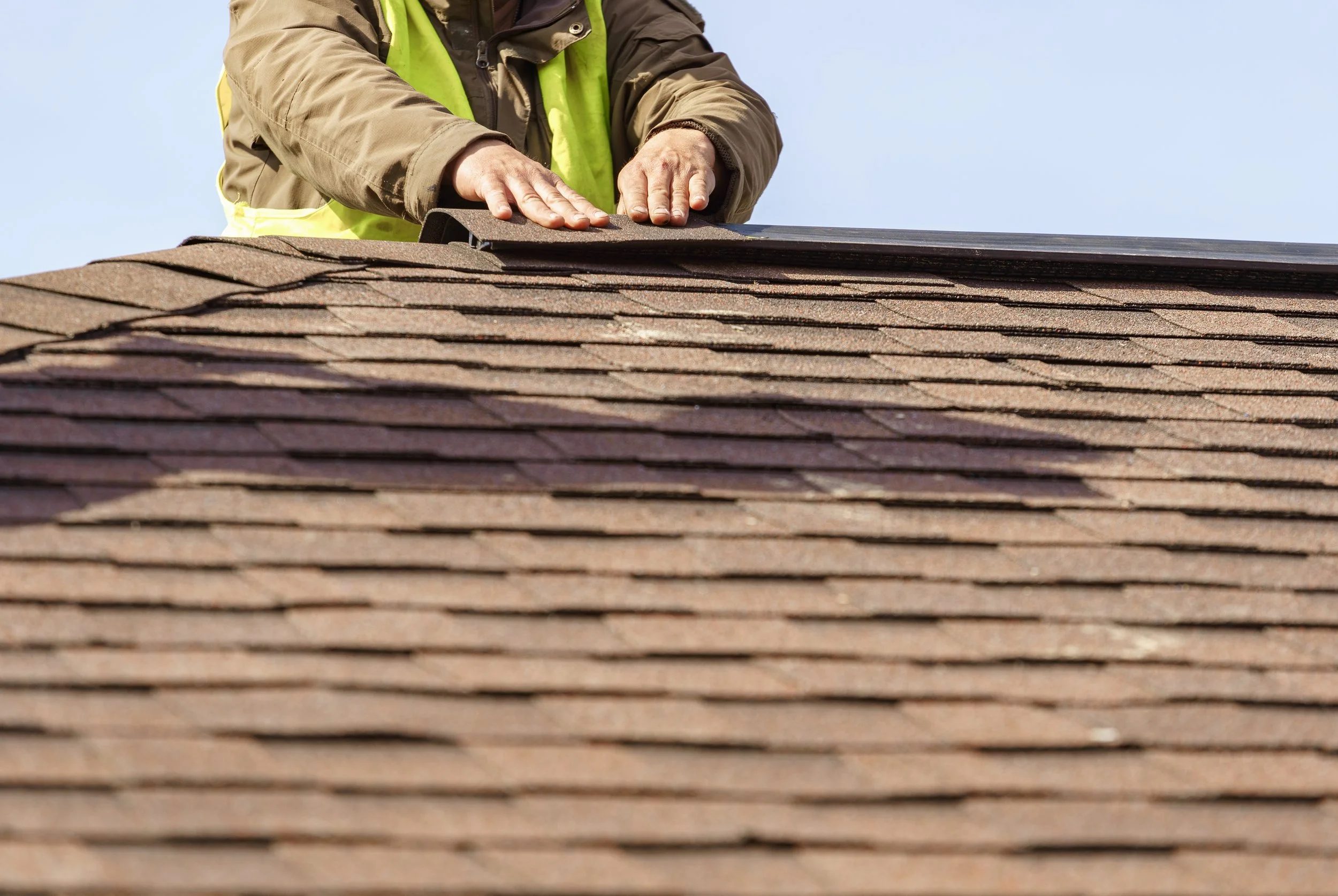 A worker repairing asphalt shingles on a roof with a sky background.