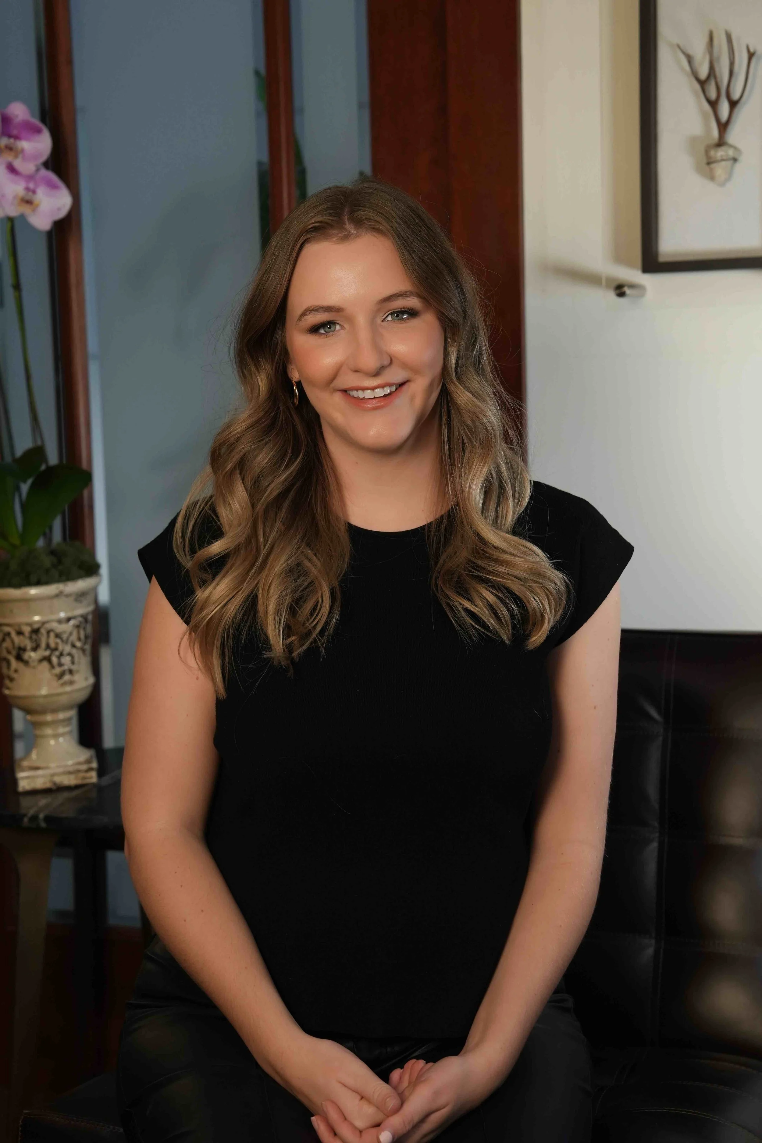 A woman with wavy, shoulder-length brown hair, wearing a black top, smiling and sitting in a room with plants and artwork.
