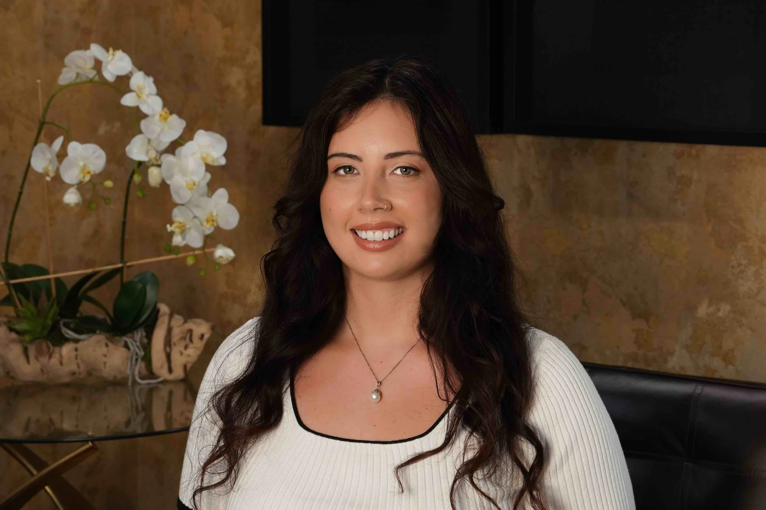 A woman with long dark wavy hair, smiling, wearing a white top with black trim, sitting indoors near a table with a white orchid plant, against a textured brown wall.