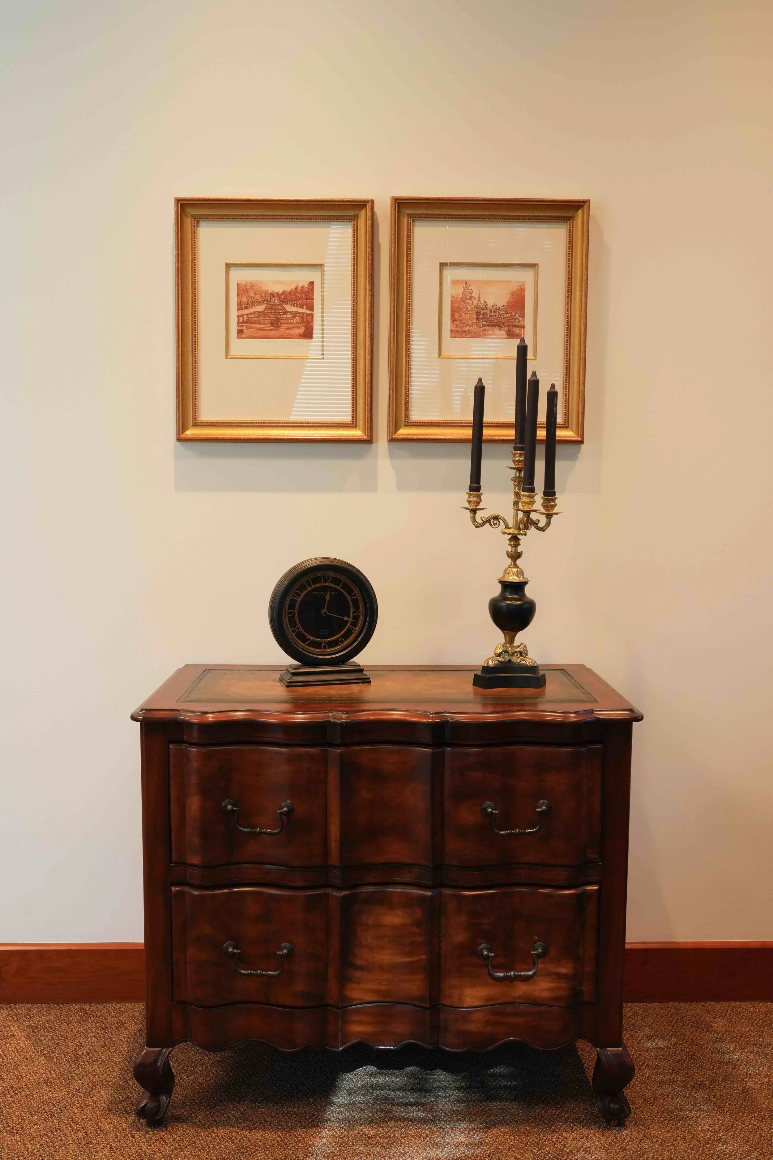 A wooden dresser with six drawers sitting against a cream-colored wall, topped with a black clock and a brass candelabrum with five black candles, above two framed pictures of cityscapes.