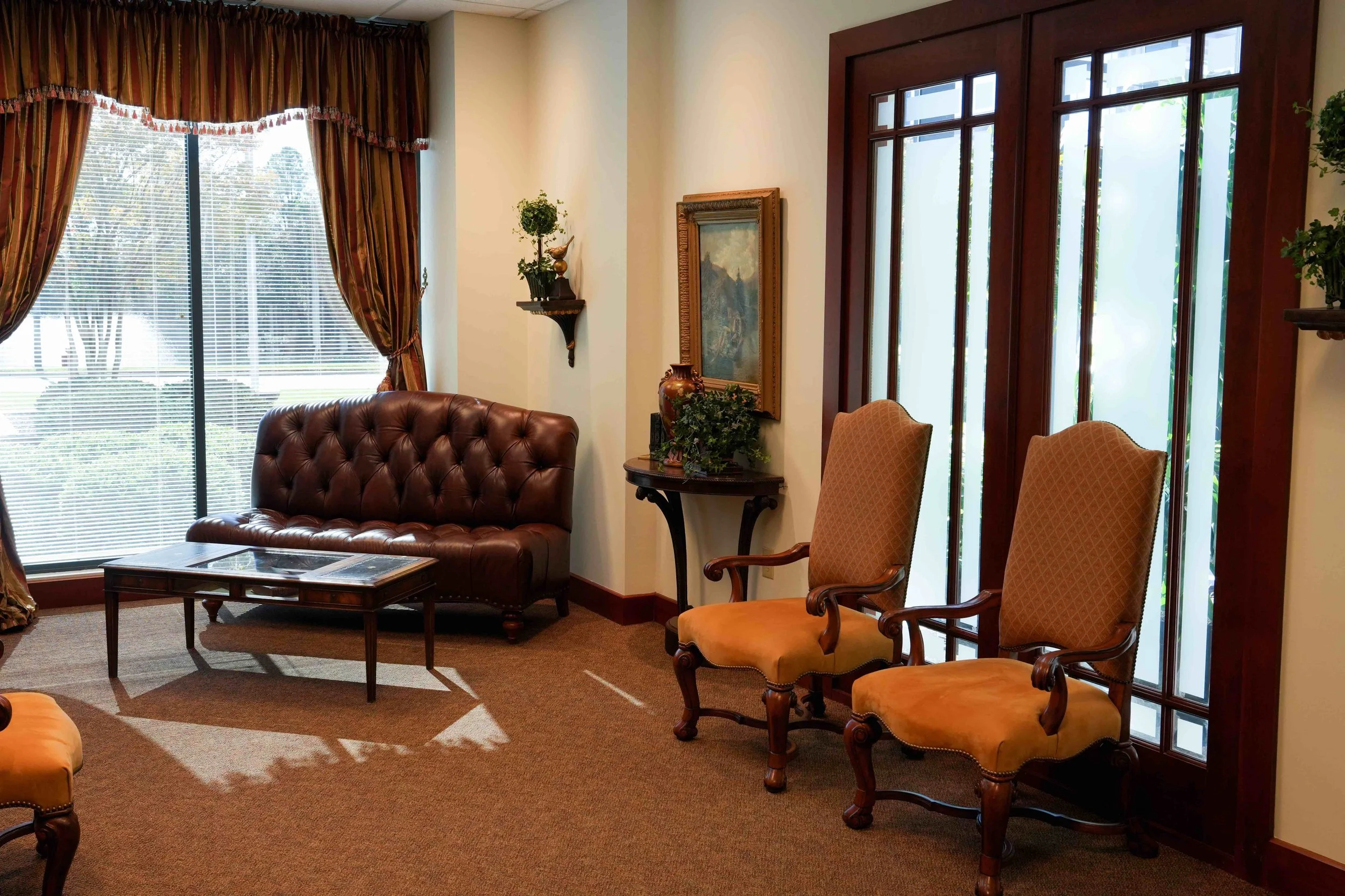 Surgical waiting room with a large window with curtains, brown leather tufted sofa, two wooden armchairs with upholstered seats, a glass-topped coffee table, decorative plant and paintings, and a door with glass panels.