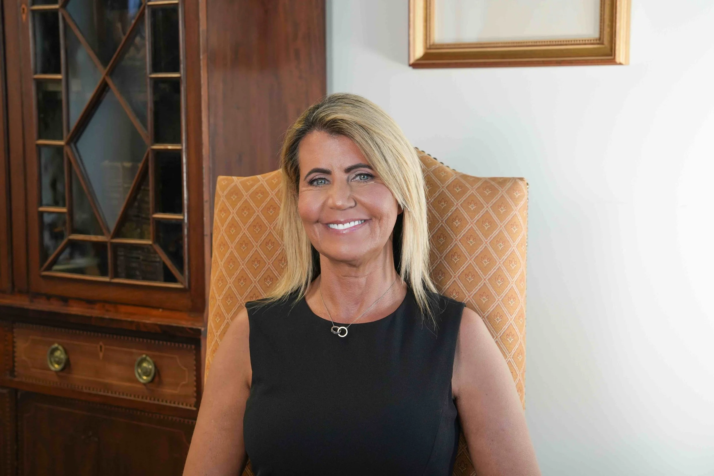 A smiling woman with blonde hair sitting on an orange patterned chair in a room with wooden furniture and a white wall.