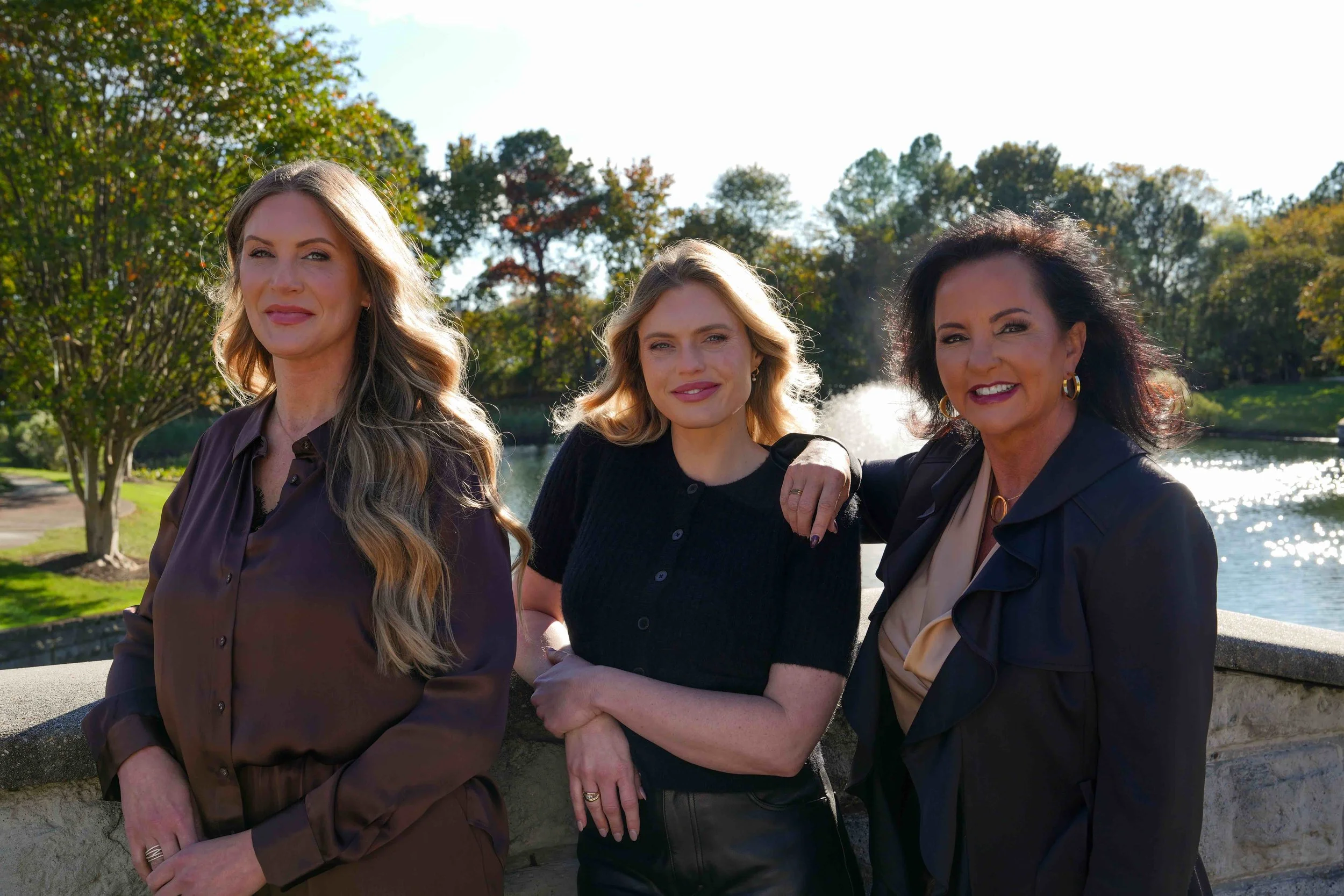 Three women standing outdoors near a body of water with trees in the background. They are smiling and dressed in dark clothing.