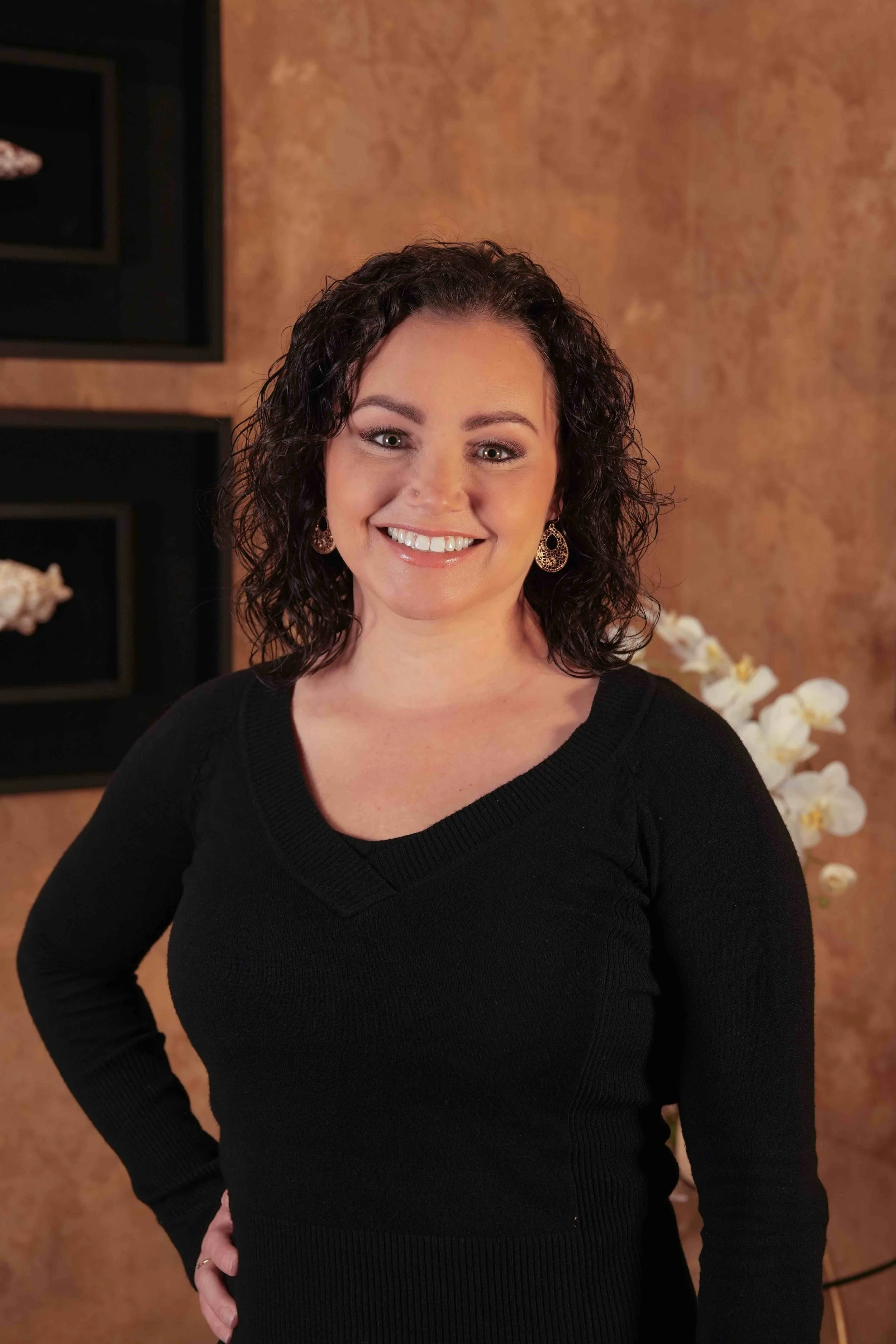A woman with curly dark hair, wearing a black sweater and earrings, smiling and posing indoors with a warm-toned wall and white flowers in the background.