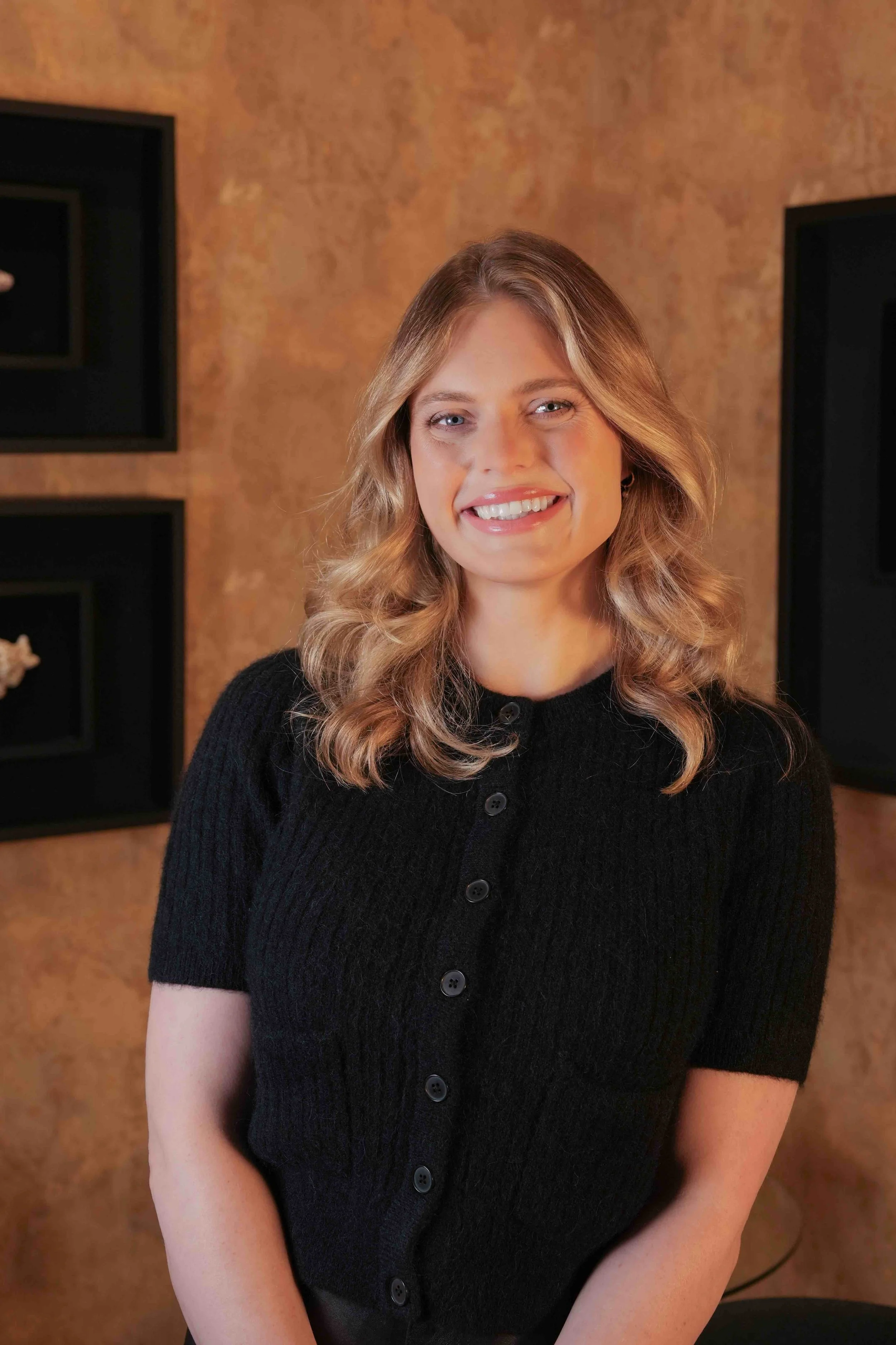 A woman with blonde, wavy hair smiling in front of a textured brown wall with black shadow boxes.