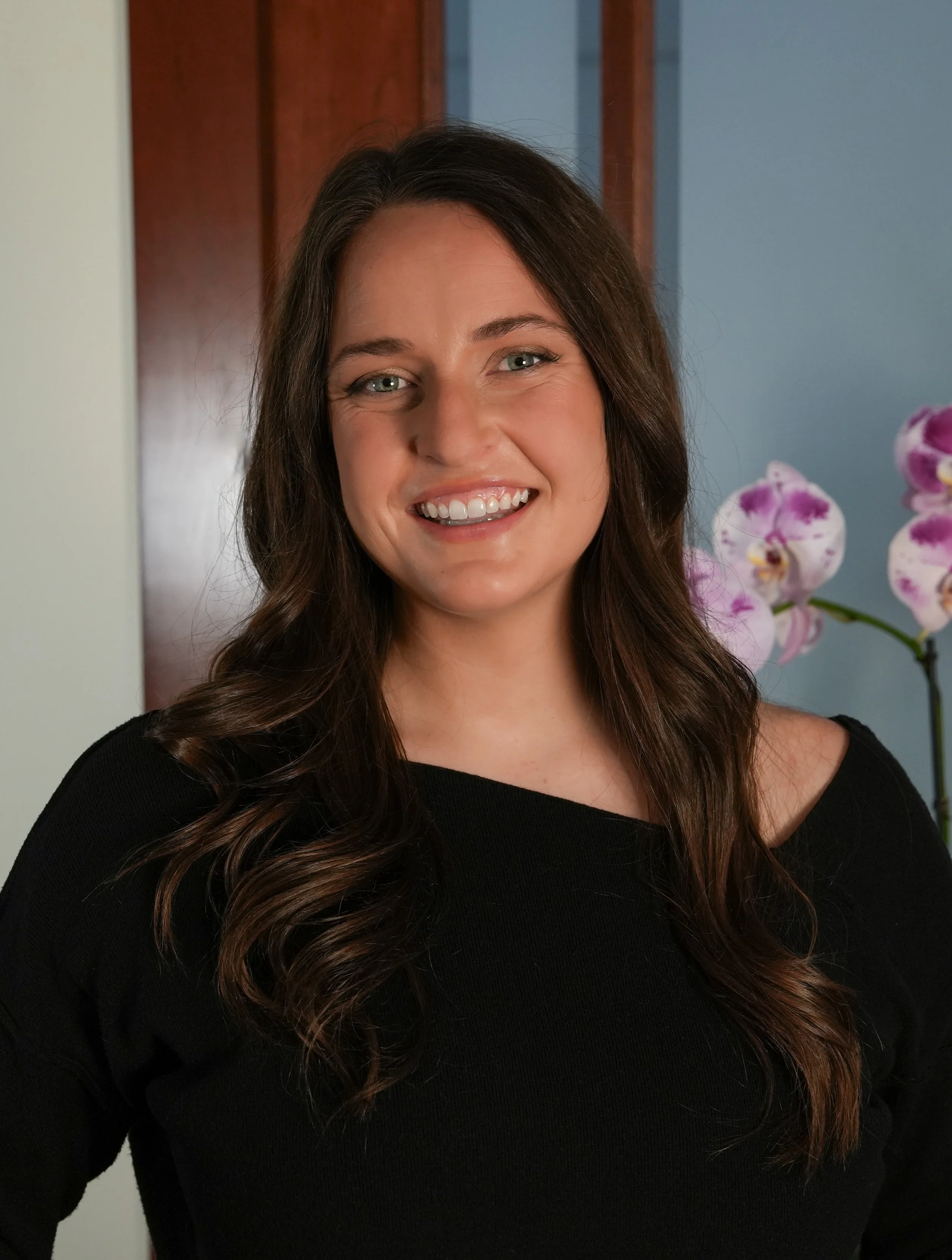 Smiling woman with long wavy brown hair wearing a black top, standing indoors near a wooden door and purple orchid flowers.