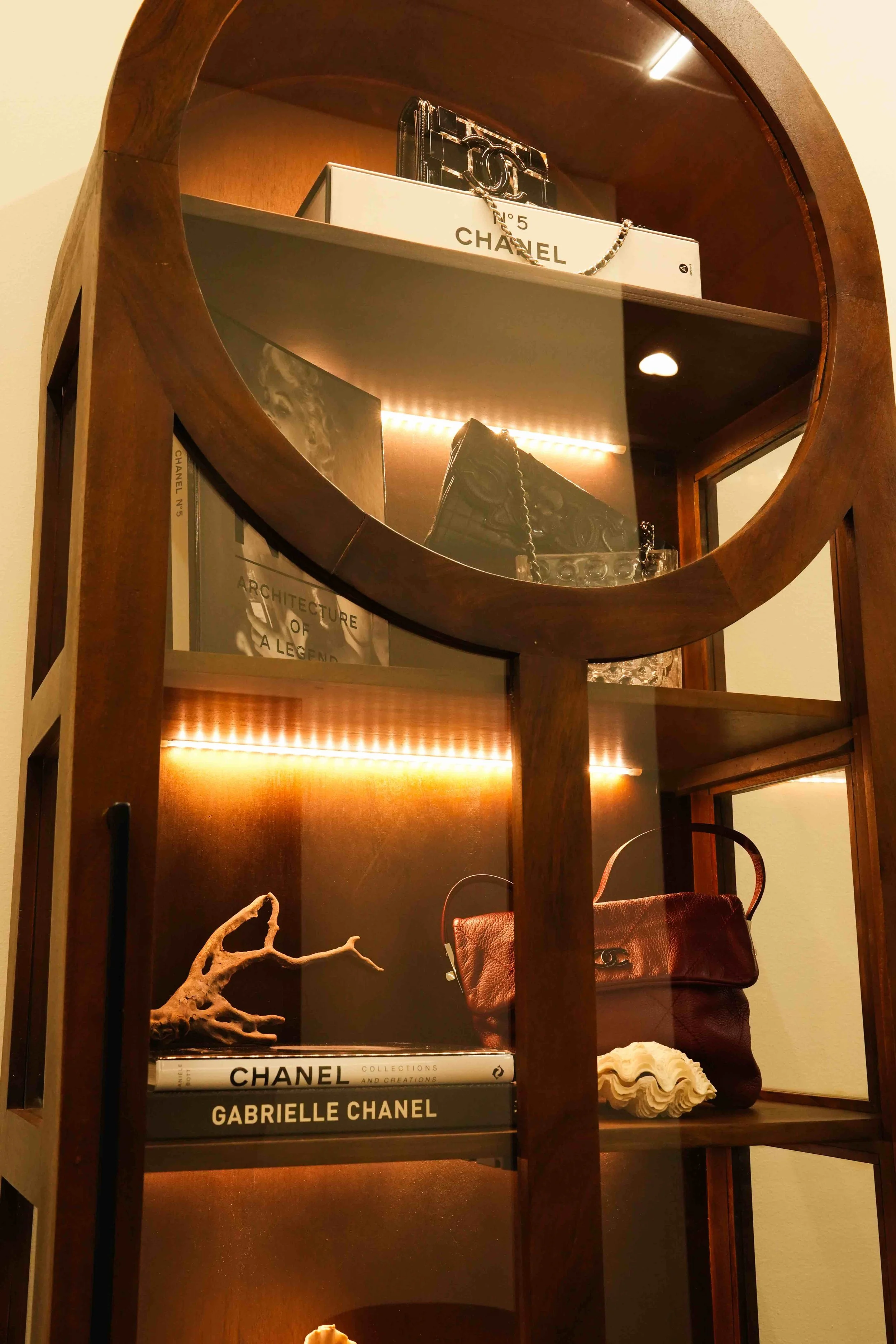A wooden shelving unit with a circular mirror, displaying fashion and design books, a small coral sculpture, a red handbag, and a white shell.