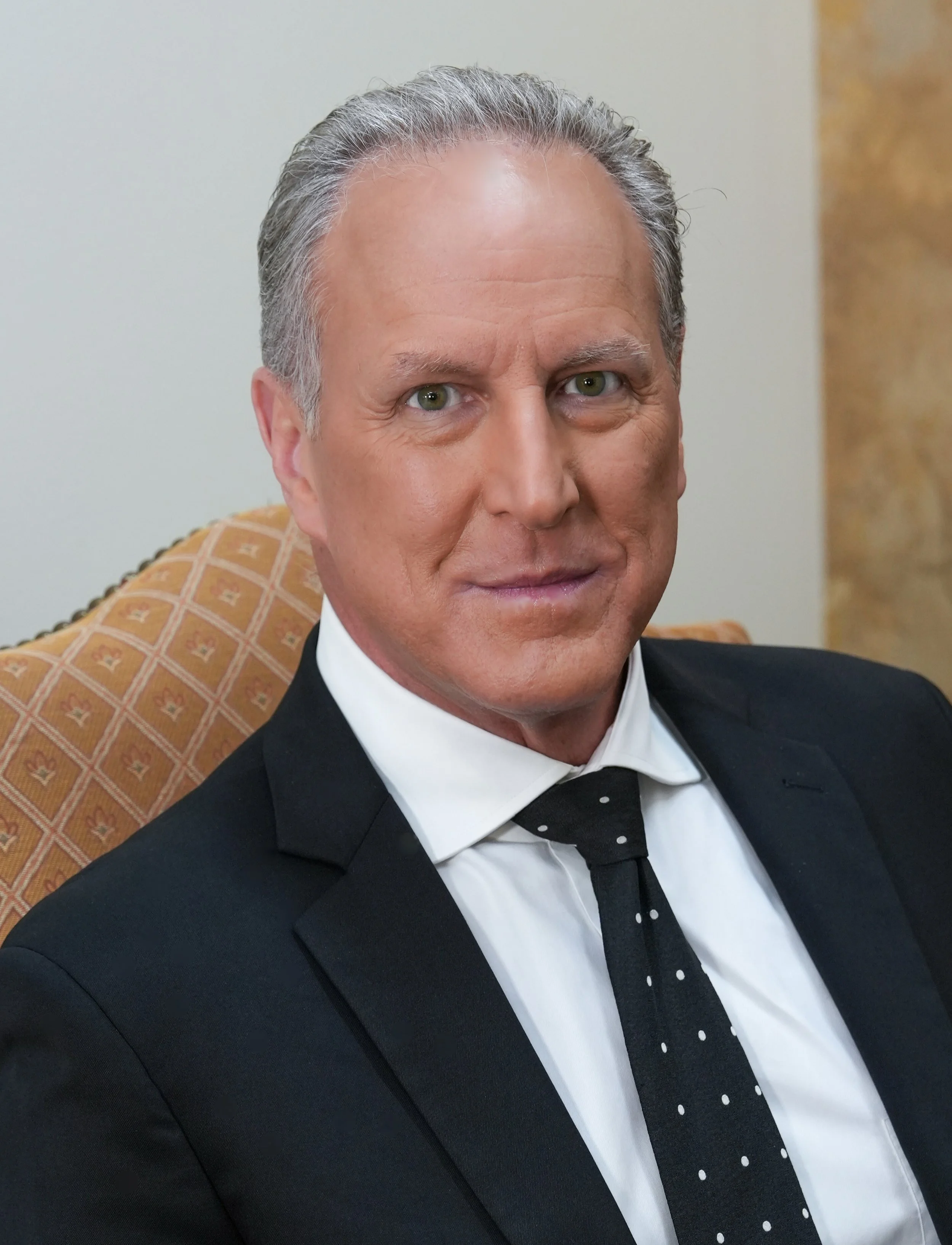 A middle-aged man with greying hair and blue eyes, wearing a black suit, white shirt, and black tie with white polka dots, sitting in a patterned orange and beige chair against a neutral background.