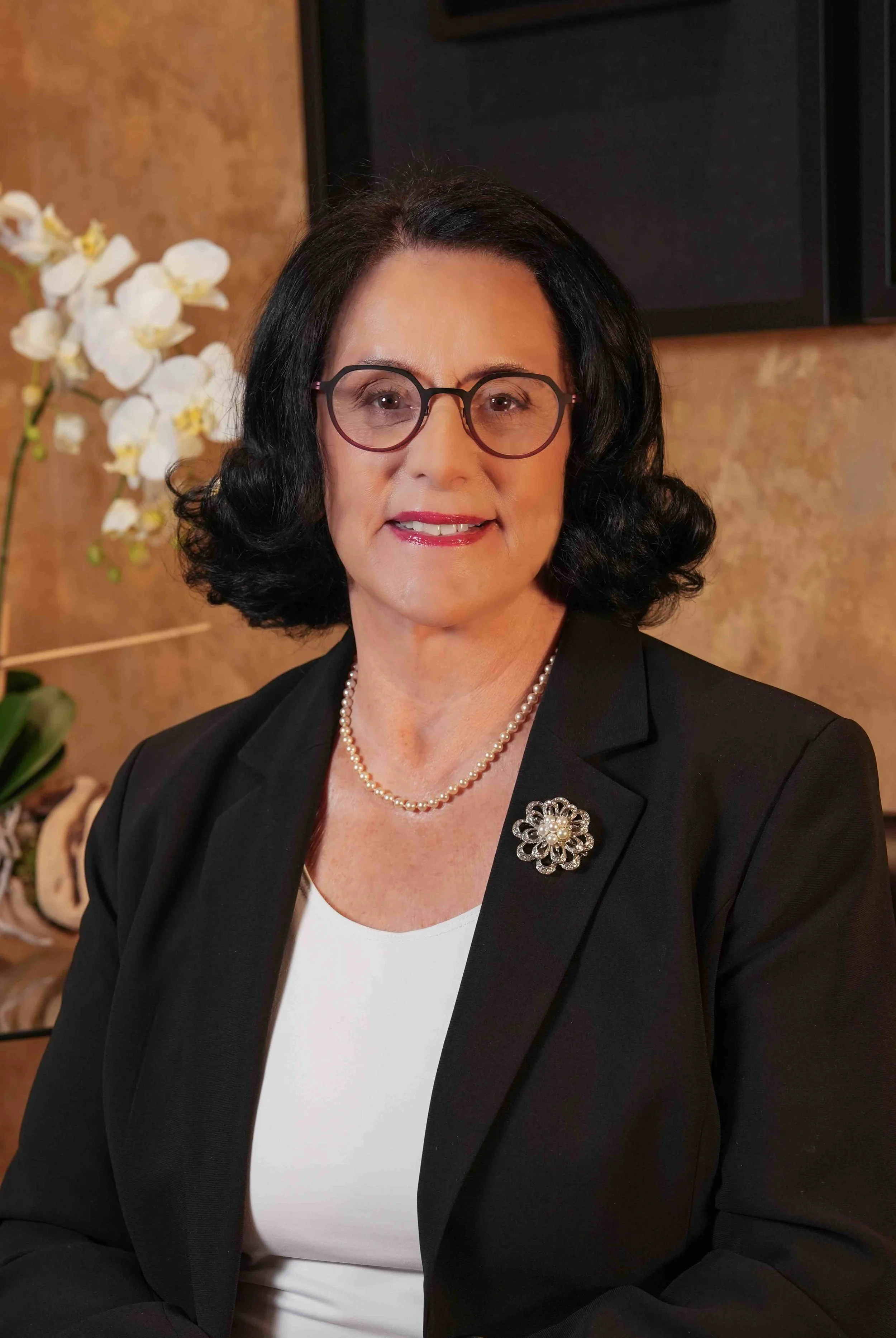 A woman with black hair, wearing glasses, a pearl necklace, and a black blazer with a decorative brooch, sitting indoors near a flower arrangement and a wall with artwork.