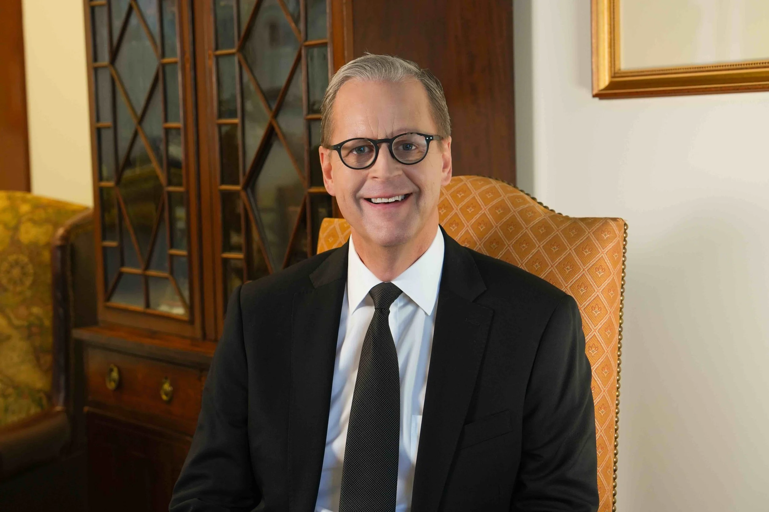 A smiling man wearing glasses, a black suit, white shirt, and black tie, sitting in an orange patterned armchair in a room with wood furniture and decorative framed mirrors.