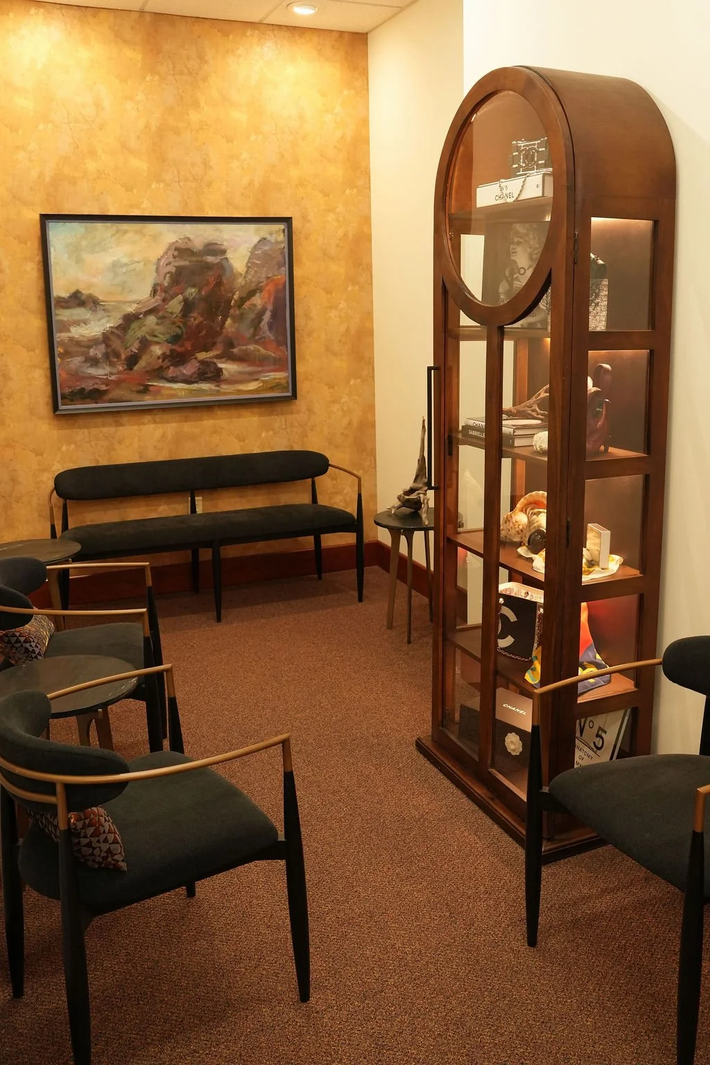 A cozy waiting room with a black sofa, chairs, a large abstract painting on a textured yellow wall, and a wooden display cabinet with decorative items and books inside.