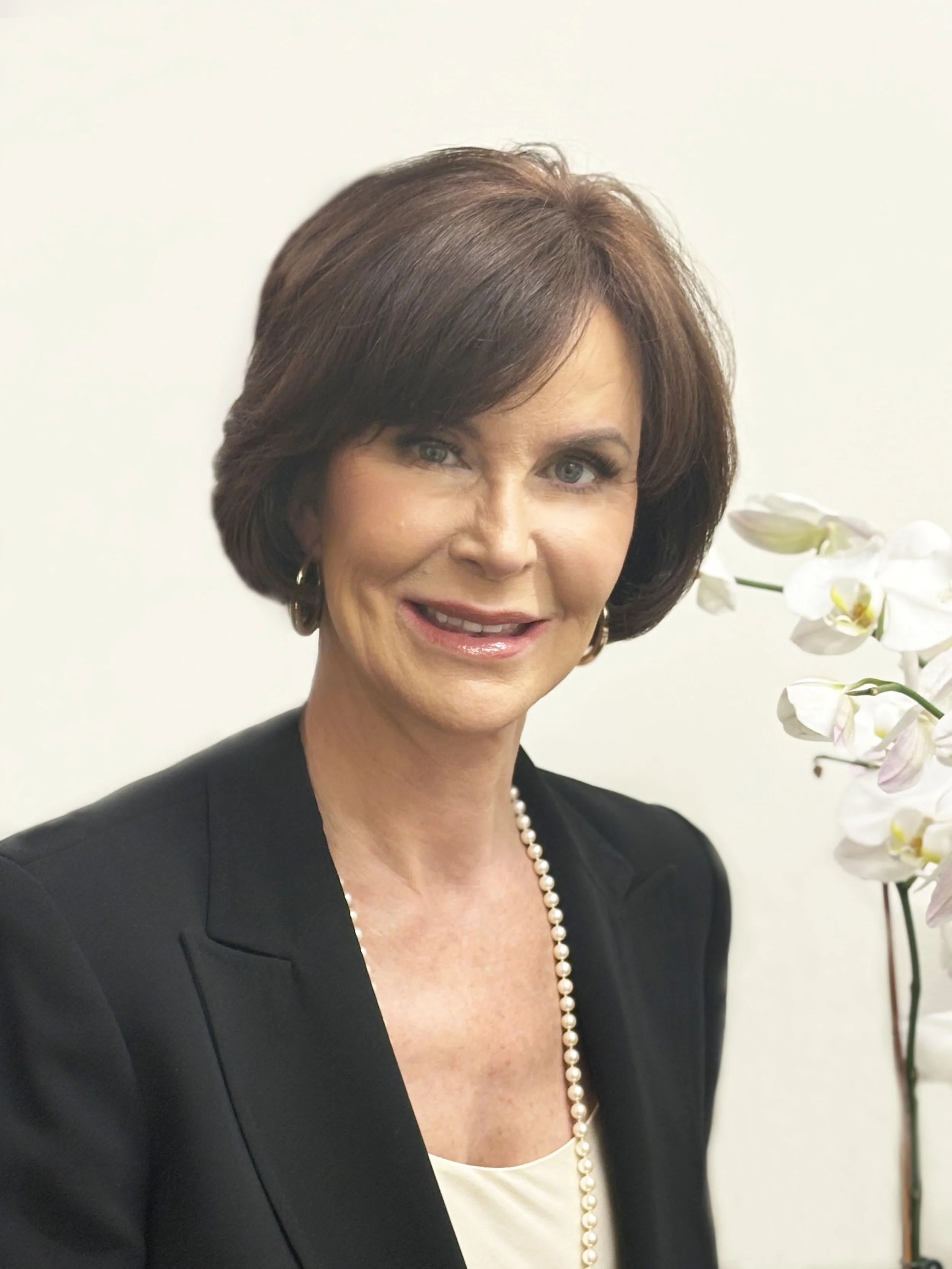 A woman with short brown hair and light skin, wearing a black blazer, a pearl necklace, and earrings, smiling at the camera with a white background and white orchids to her right.