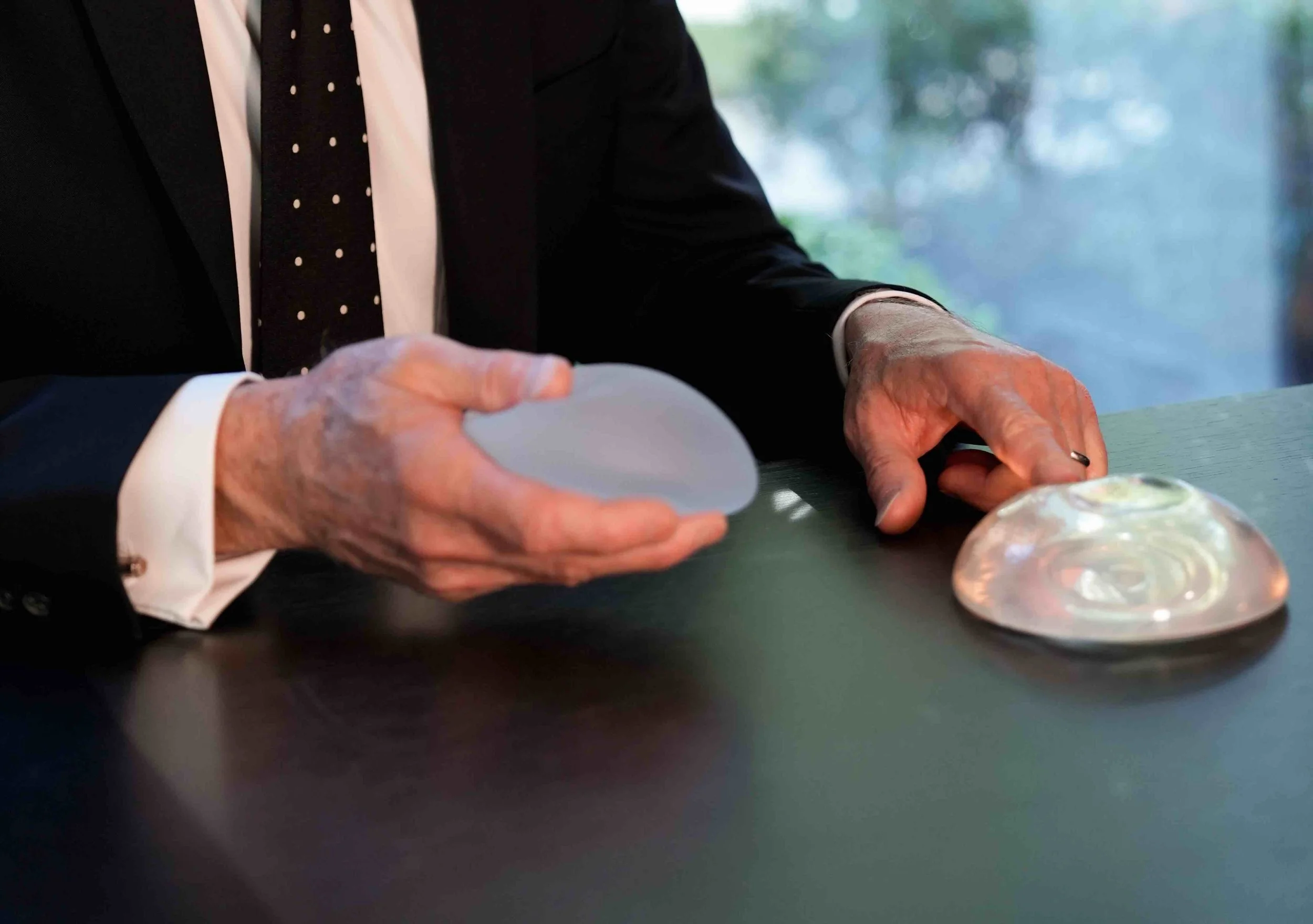 A man in a black suit with a white shirt and a black polka-dotted tie is sitting at a desk, holding a round breast implant in one hand and pointing to another on the table with the other hand.
