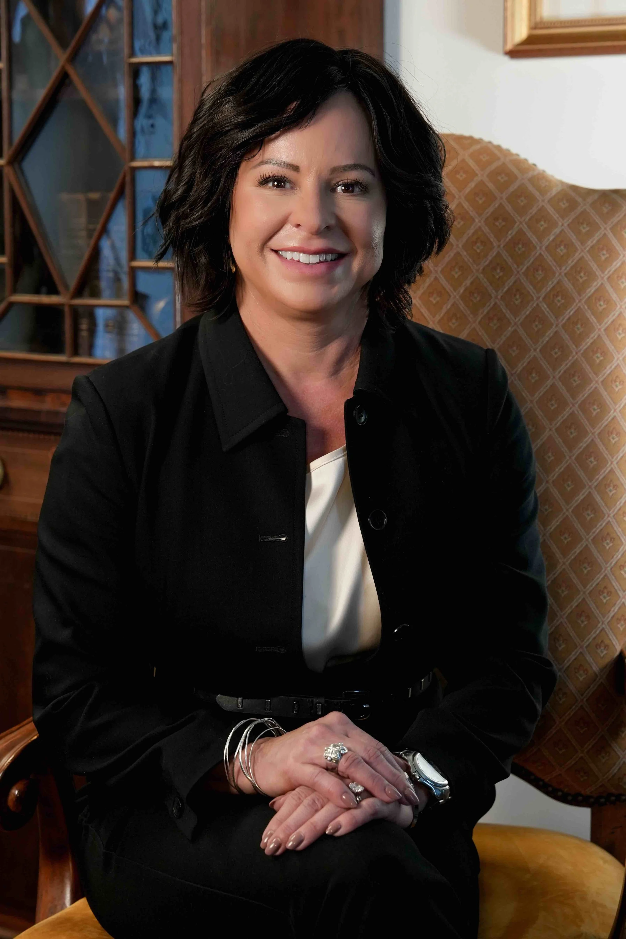 A woman with short black hair smiling, sitting in a wooden chair with a patterned upholstery. She is wearing a black jacket over a cream top, with rings, bracelets, and a watch on her left hand, and a ring on her right hand. The background includes a wooden cabinet with blue glass doors and a framed picture on the wall.