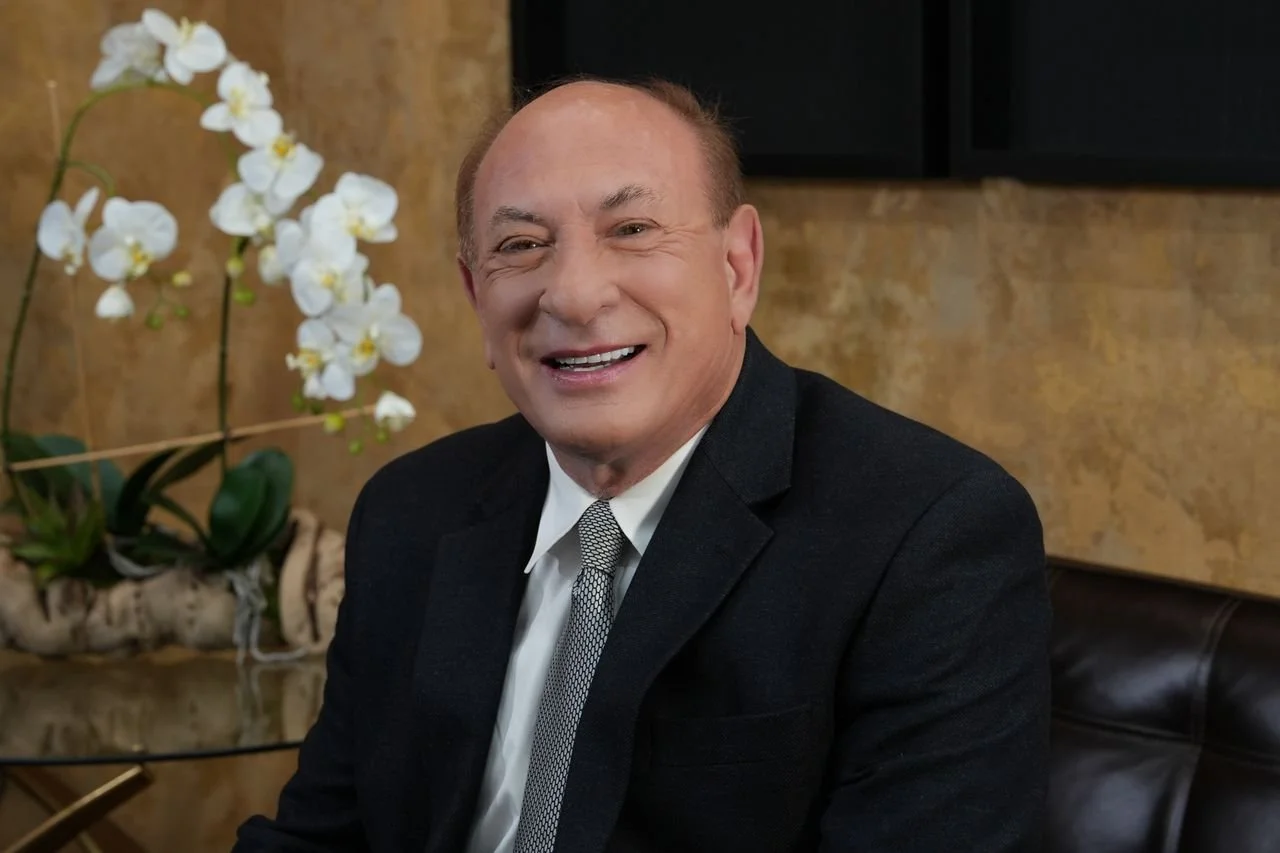 Smiling man in a black suit and white shirt sitting on a leather chair with a white orchid plant in the background.