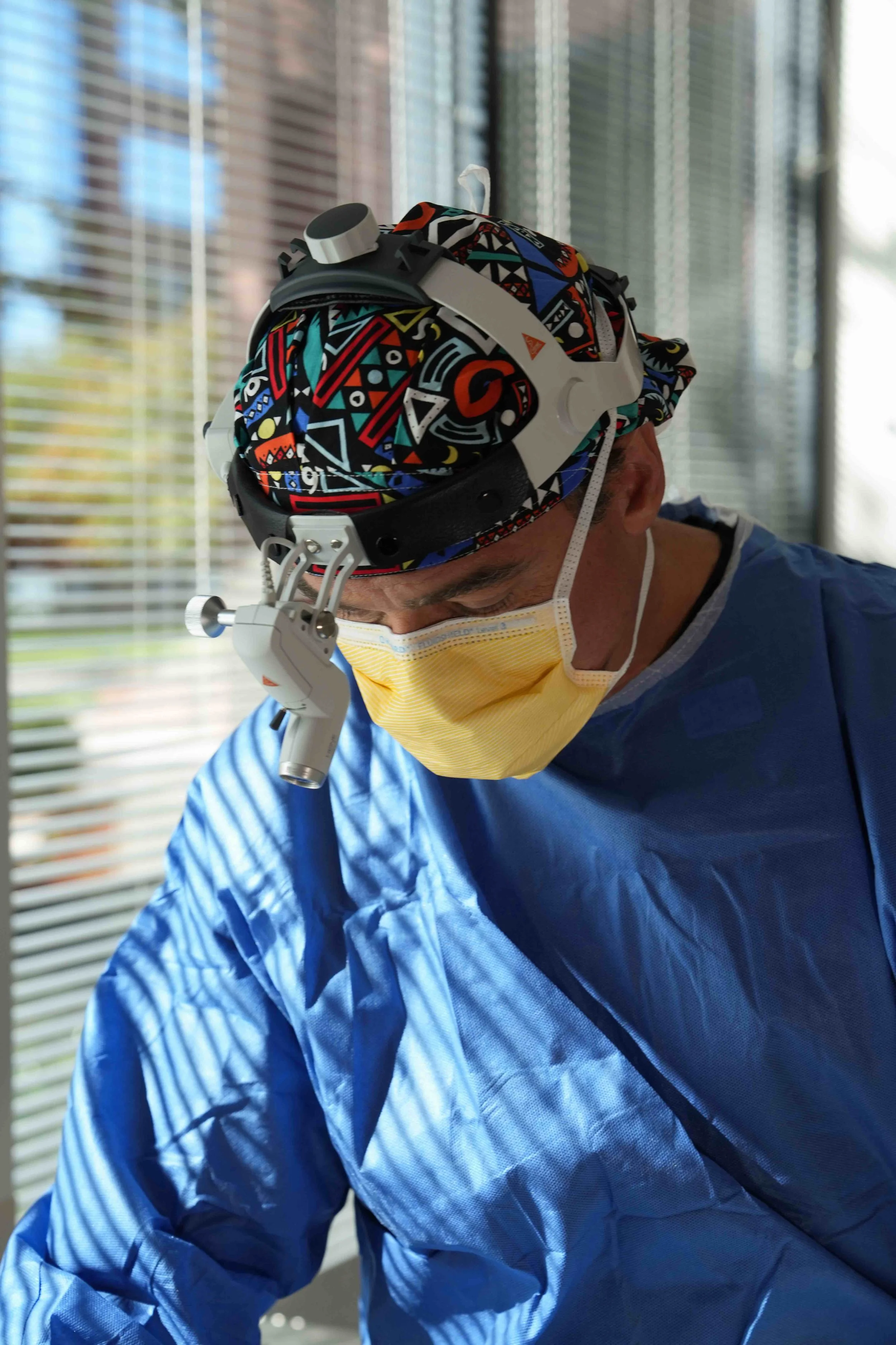 A healthcare professional wearing a colorful patterned cap, a yellow face mask, and blue medical gown, looking down with a headlamp on his forehead in a clinical setting with window blinds in the background.