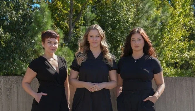 Three women standing outdoors in front of a concrete wall with green trees in the background