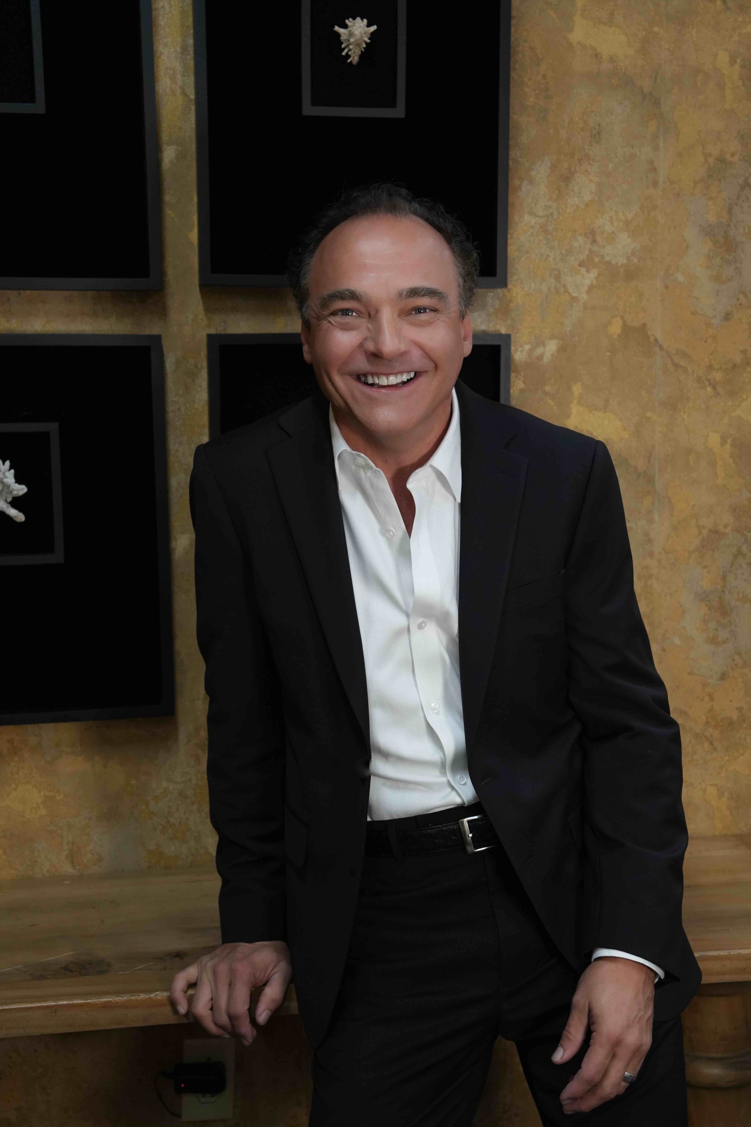 A man in a black suit and white shirt smiling and leaning against a wooden table, with a textured yellow wall and black-framed artwork behind him.