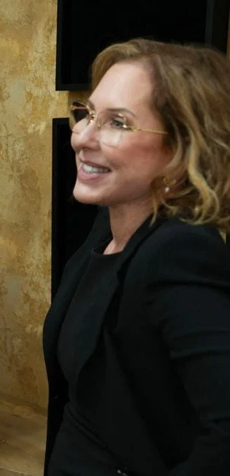 A woman with light brown, shoulder-length, wavy hair wearing glasses and a black blazer, smiling and looking to the right.