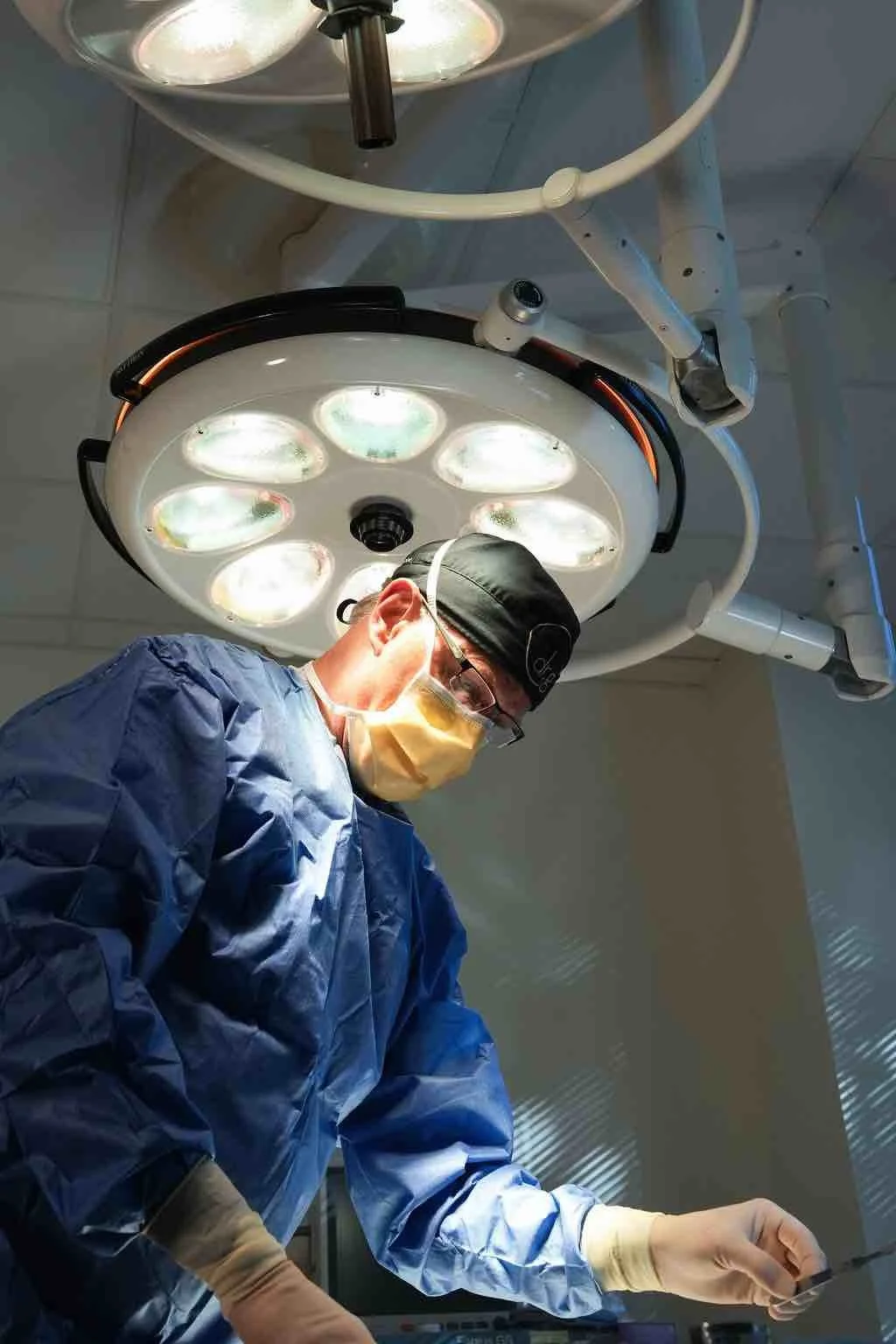 A surgeon in scrubs, mask, and glasses operating under bright surgical lights in an operating room.