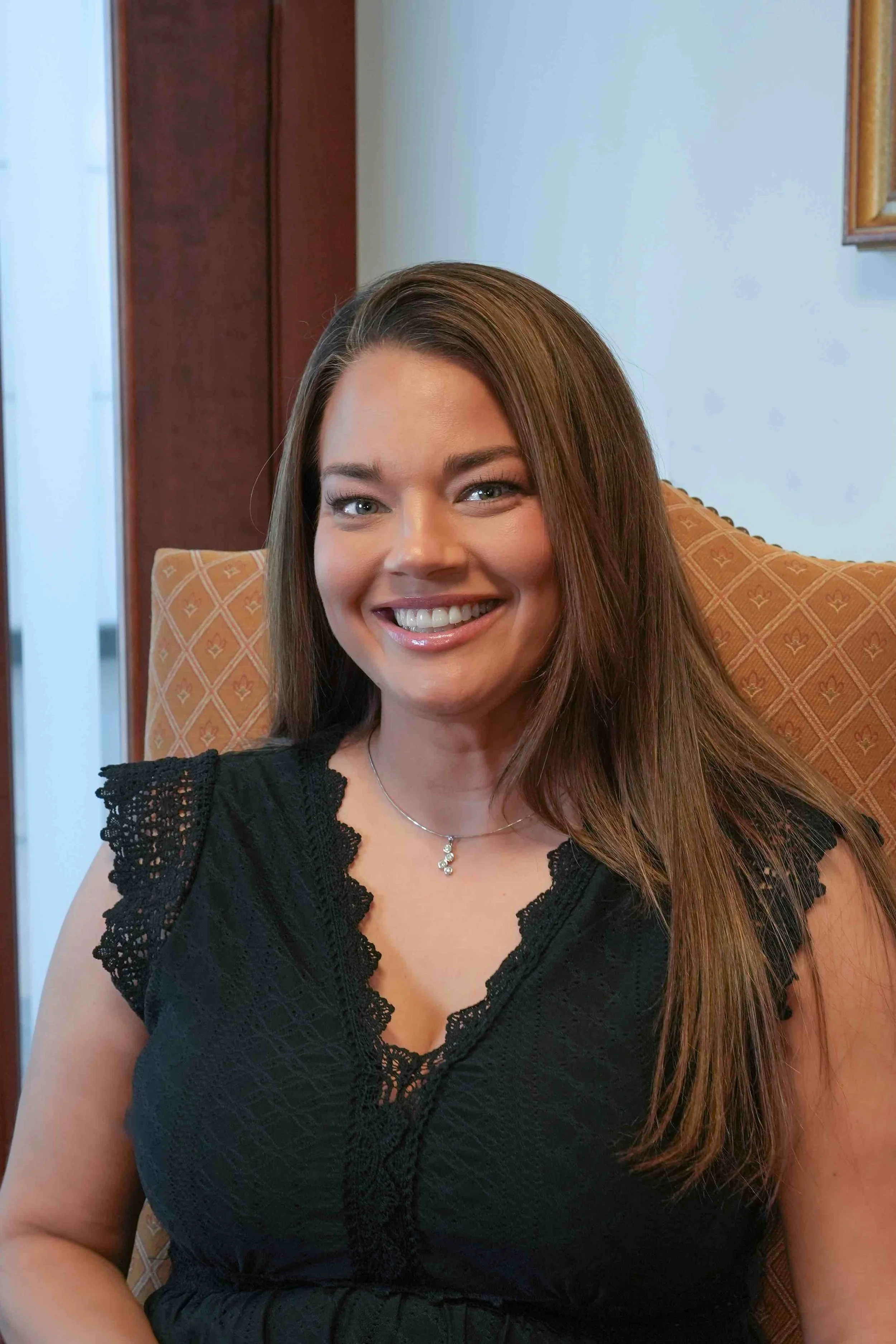 A smiling woman with long brown hair wearing a black lace dress and a silver necklace, sitting on an orange patterned chair in a room with a wooden door and painting.