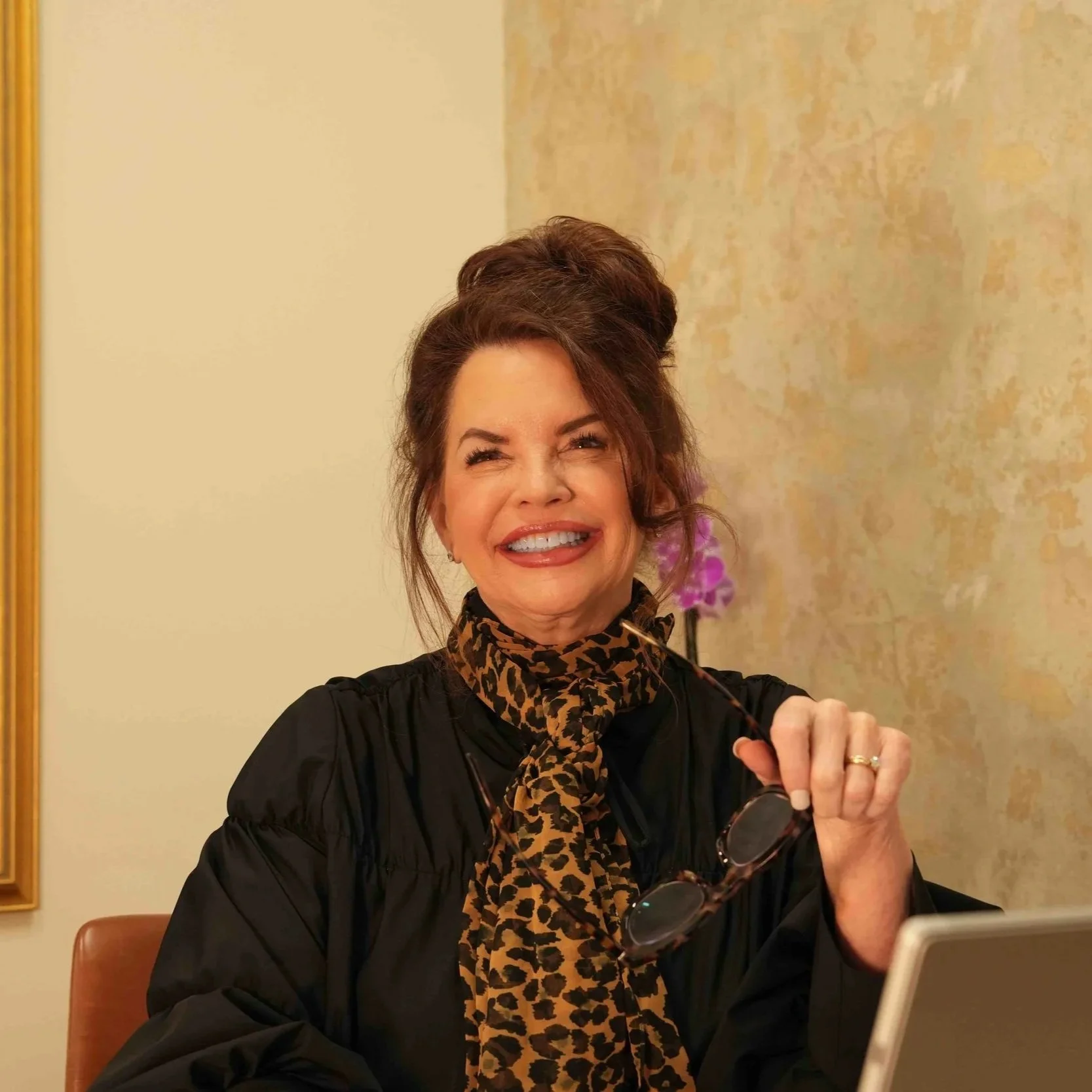 A smiling woman with brown hair styled in an updo, wearing a black top and a leopard print scarf, holding glasses, sitting at a desk in an office setting.
