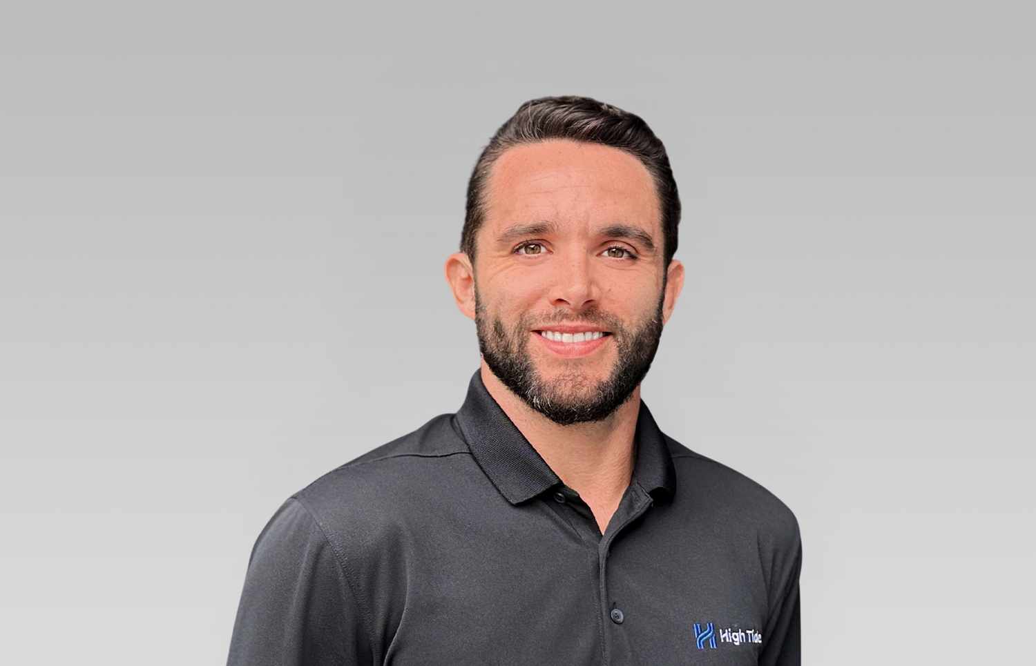 A smiling man wearing a black collared shirt with a logo that says 'High Tide' on the left chest, standing against a plain grey background.