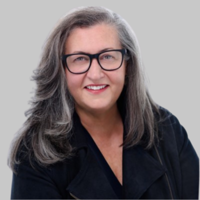 A woman with long, gray hair wearing glasses and a dark purple top, posing against a plain, light-colored background.