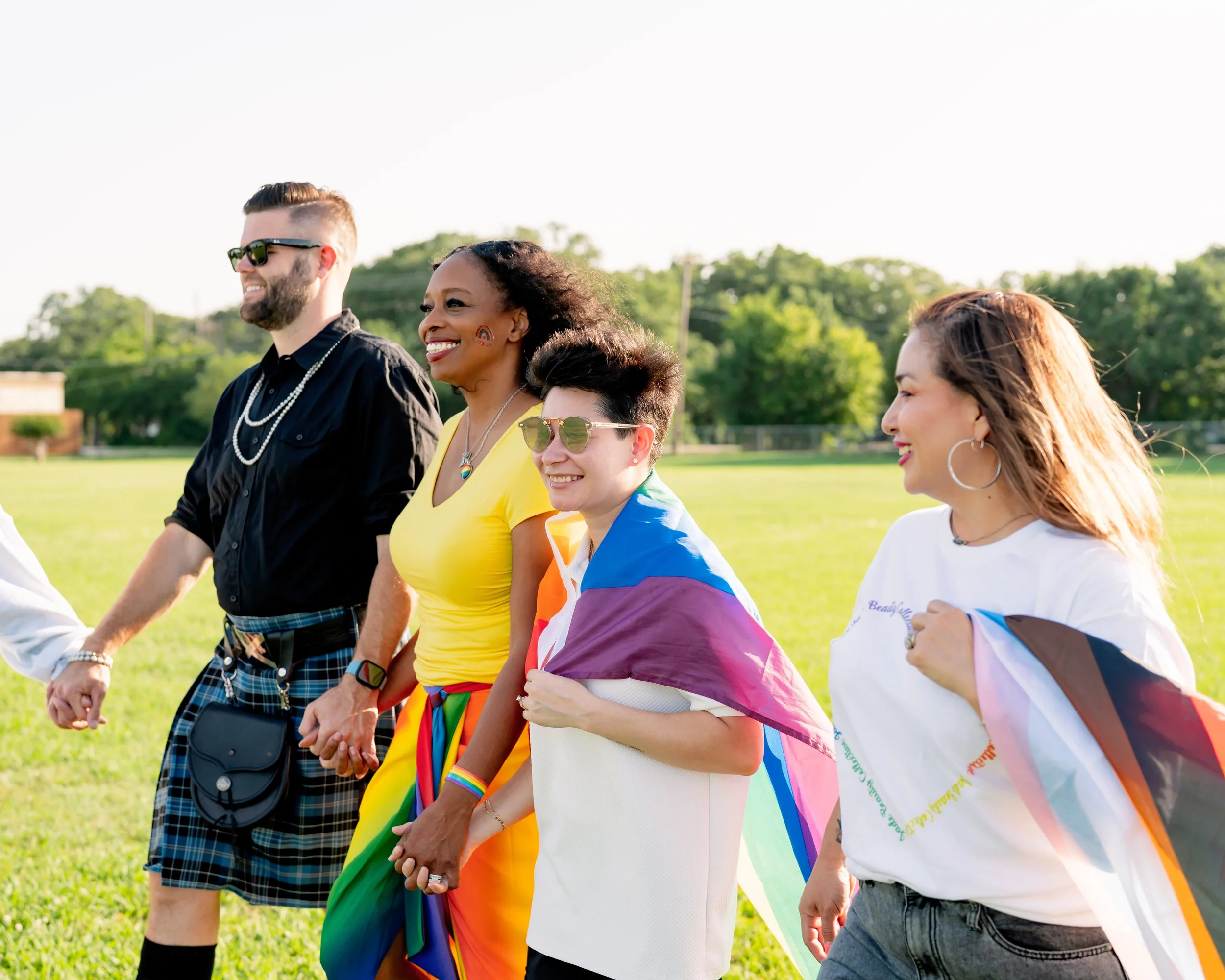 Group of diverse people holding hands and smiling outdoors, celebrating LGBTQ+ pride with rainbow flags and colorful clothing.