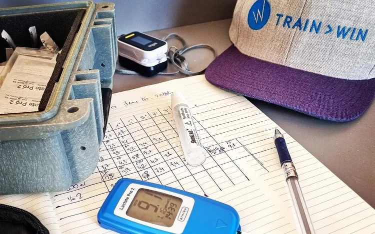 A Lactate Pro 2 meter on a handwritten lactate step-test sheet next to a Train2Win cap