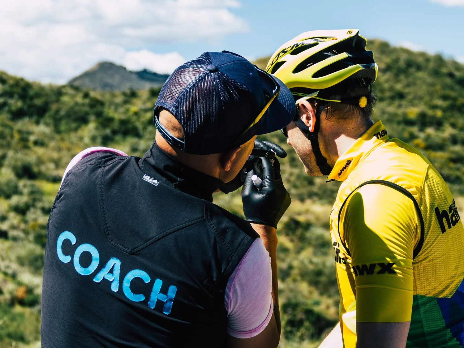 Jānis Mūsiņš, in a vest marked COACH, takes a lactate blood sample from a rider during a field test on a hillside