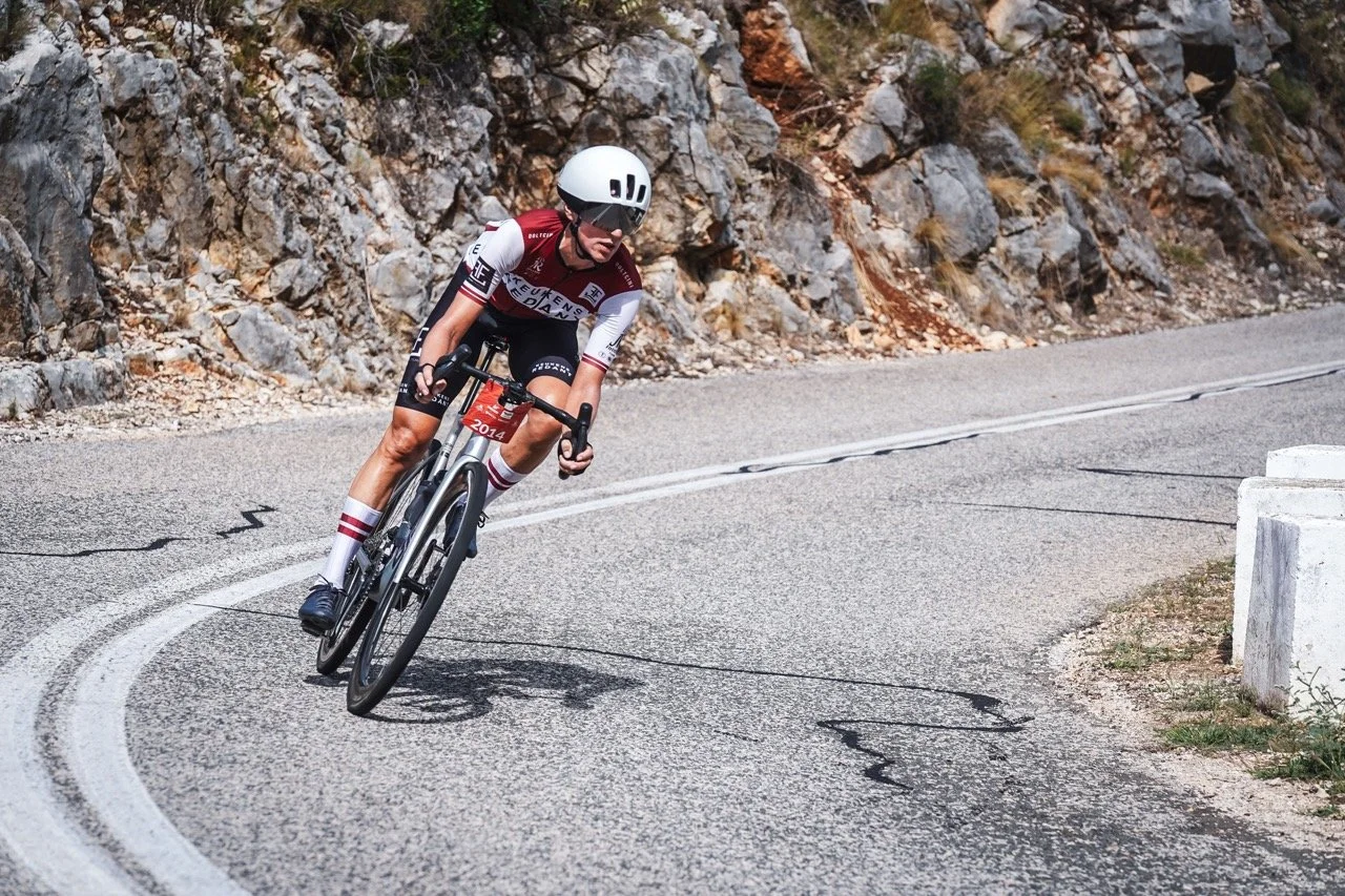 A cyclist with a race number leans into a hairpin bend on a mountain road below a rock face